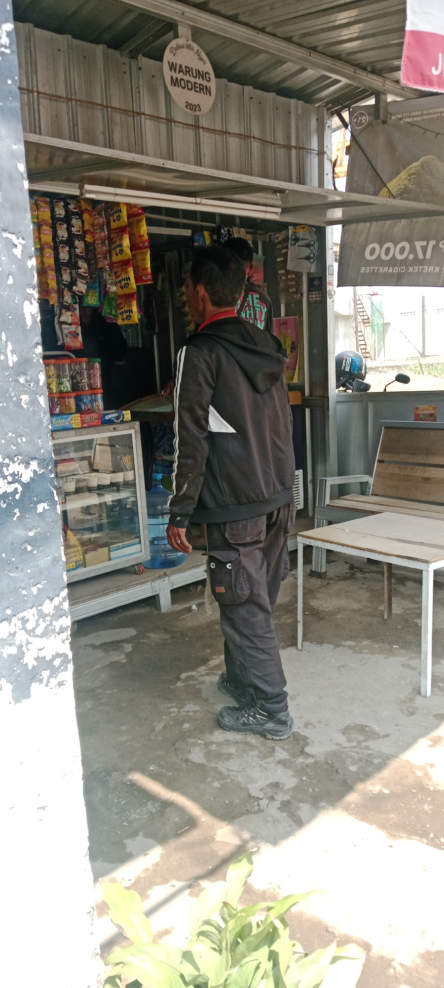 In the city of Purwomartani, Indonesia, a man stands in front of a small, open-air convenience store, identified by a circular sign reading "WARUNG MODERN 2023." The man is wearing a black hooded jacket with white stripes on the sleeves and black cargo pants, along with sturdy black boots. He is facing away from the camera, looking towards the store's interior. The store displays various packaged snacks and drinks on shelves and hooks. To the right of the store, a wooden bench and a table are visible, with a motorcycle helmet resting on a metal railing behind them. The ground is a mix of concrete and dirt. The scene is outdoors, under a corrugated metal roof that provides shade.