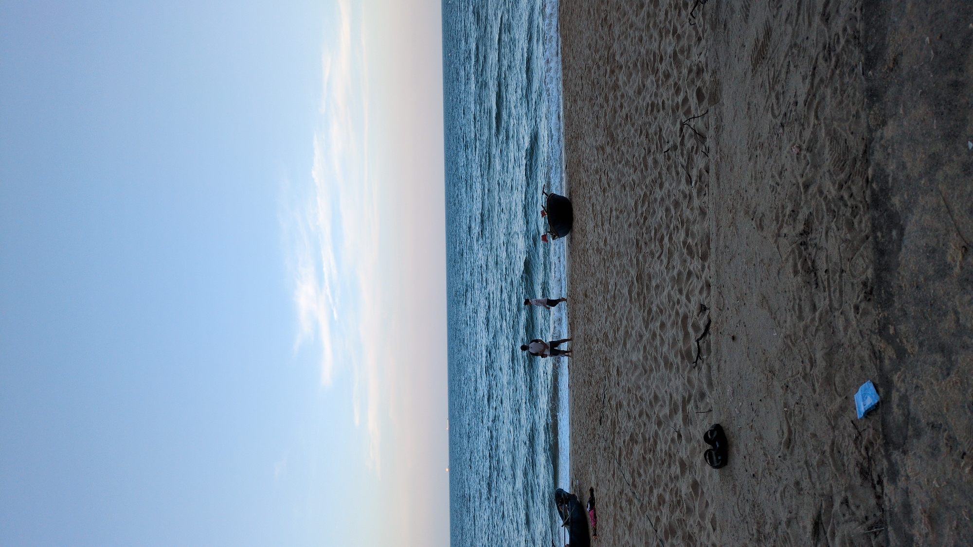The image shows a beach scene in South Quy Nhon Ward, Vietnam. The sky is light blue with faint clouds. The ocean is deep blue with visible waves. Several people are standing at the shoreline. There are two boats on the sand near the water. A pair of black sandals and a small blue object are on the sand further inland. The sand is light brown with varied textures.