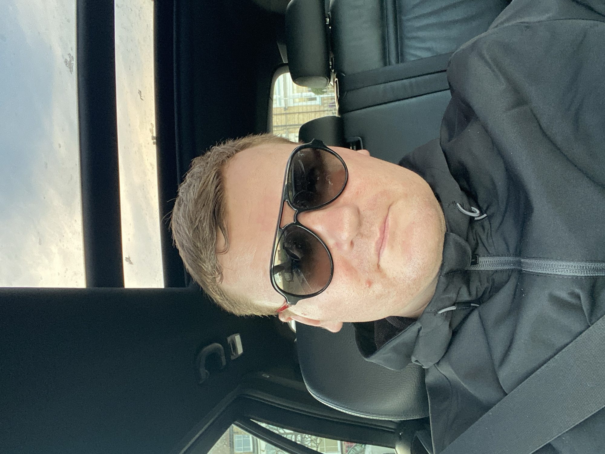 A middle-aged man wearing sunglasses and a black jacket is sitting in a vehicle. He is wearing a seatbelt. The vehicle appears to be stationary. The background shows buildings that may be located in East Suffolk, United Kingdom. The car interior is mostly black leather. The sky is overcast.