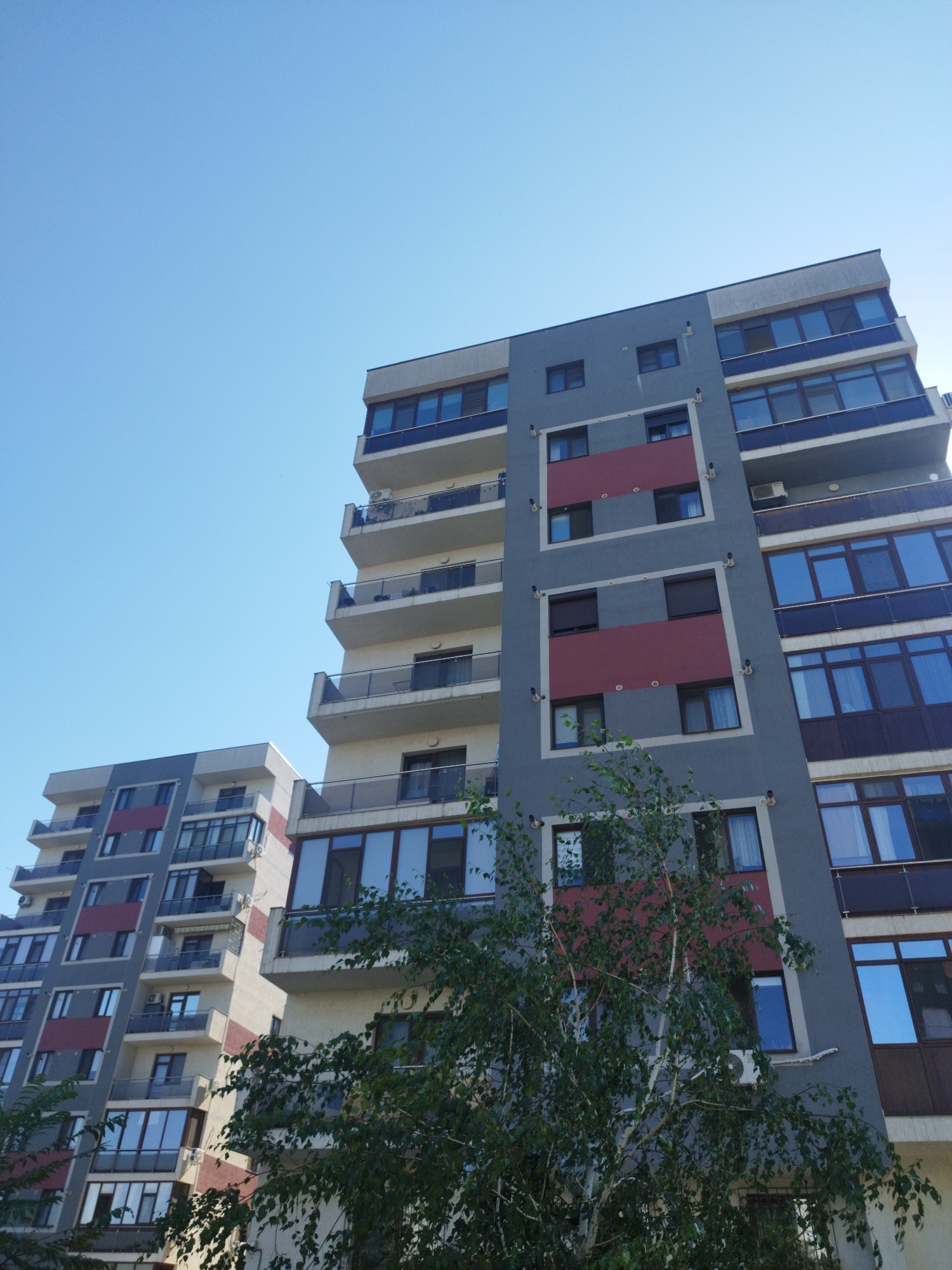 The image shows a low-angle, wide shot of two modern apartment buildings against a clear blue sky. The building on the left is shorter and has a beige and gray facade with dark-framed windows and balconies. The building on the right is taller, with a gray facade accented by rectangular maroon panels and numerous glass-enclosed balconies. Both buildings feature multiple stories with repeating window patterns and visible air conditioning units. A leafy green tree is situated in the foreground, partially obscuring the lower floors of the buildings. The architectural style is contemporary, and the scene suggests a residential area in a city. The image was taken in Bucharest, Romania.