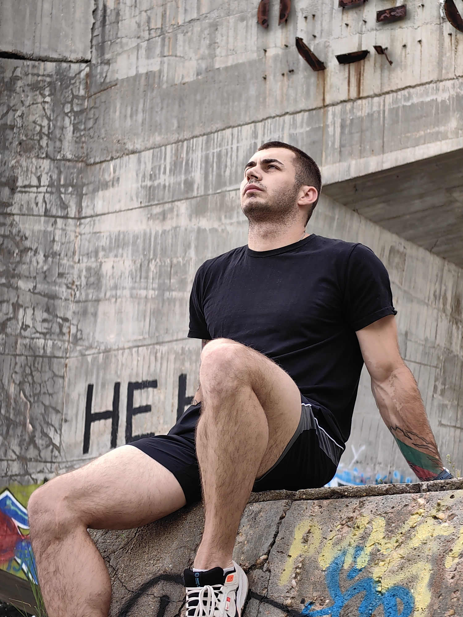 A young man sits on a concrete ledge with graffiti, situated against a large, aged concrete structure in Varna, Bulgaria. The structure features horizontal lines and vertical cracks, with rusted metal pieces protruding from its upper section. "HELP" is spray-painted in black on the concrete behind him. The man wears a black t-shirt and black shorts with a white stripe. He has short, dark hair and a beard, and his gaze is directed upwards. A tattoo is visible on his left forearm. He wears white and orange athletic shoes. The overall setting appears to be urban and slightly derelict.