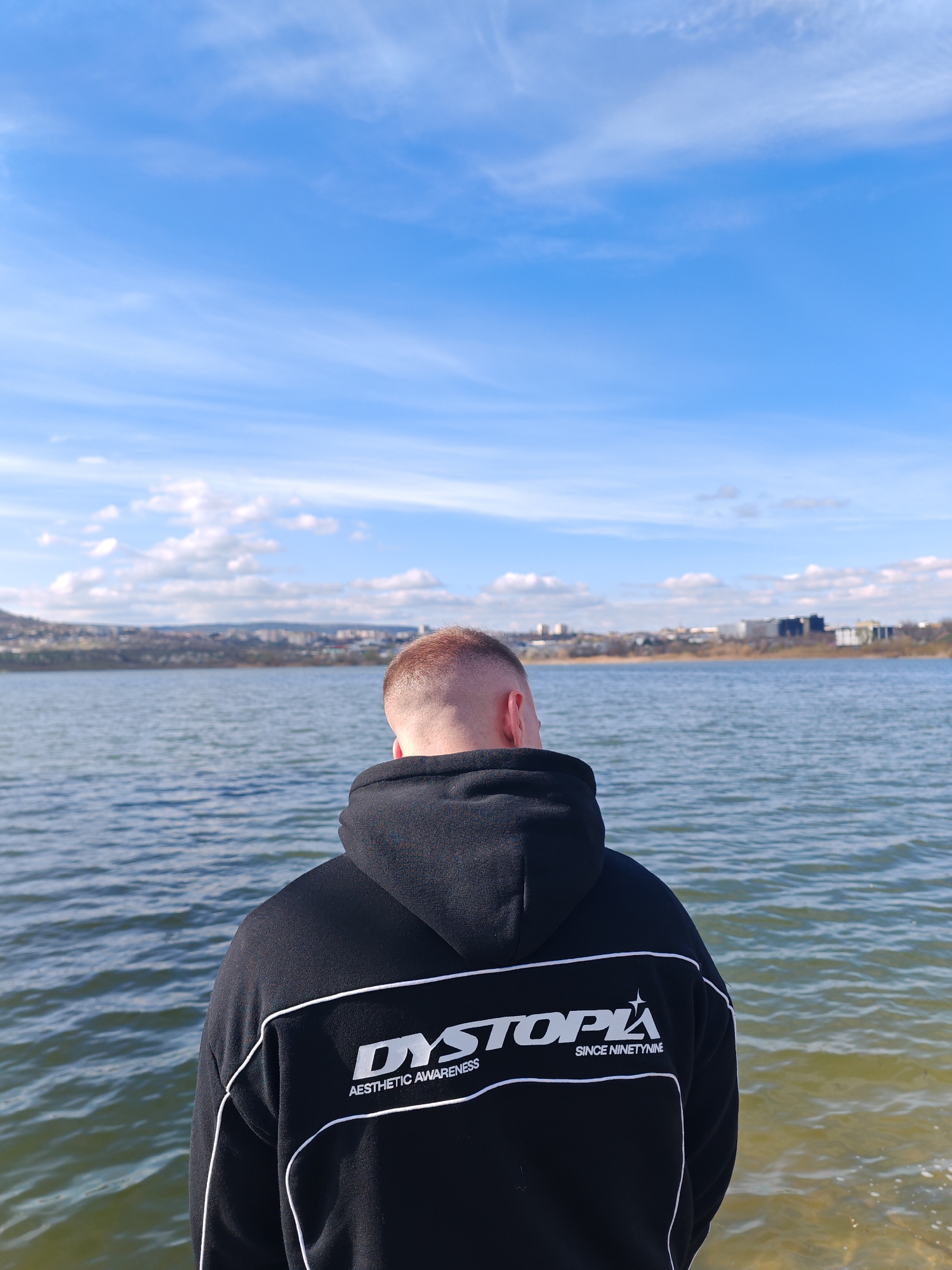 A medium shot depicts a person with a shaved head and wearing a black hoodie with white trim and the word "DYSTOPIA" printed on the back, standing with their back to the viewer near a body of water on a sunny day. The water stretches out to a distant shoreline, where buildings of varying heights and colors can be seen. The sky is a bright blue with scattered white clouds. The person is on the shore of the artificial Lake Kyosha in Shumen, Bulgaria, and looking towards the skyline of Shumen. The ground at the very bottom of the image appears to be sandy or pebbly. The hoodie has the phrase "AESTHETIC AWARENESS" and "SINCE NINETYNINE" printed underneath the word DYSTOPIA.