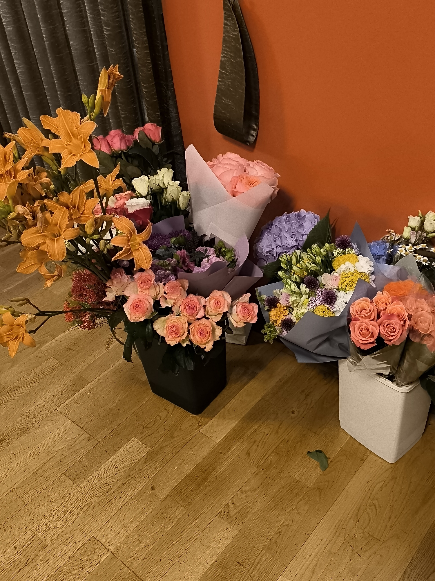 The image depicts a collection of bouquets of flowers arranged on a wooden floor next to a textured grey curtain and an orange wall. Several bouquets are in various stages of arrangement, some wrapped in paper and others in containers. The flowers are a mix of colors and types, including orange lilies, pink roses, light pink roses, white roses, purple hydrangeas, and yellow and white mixed flowers. One bouquet of orange lilies is prominently displayed on the left side of the image. To the right, there are bouquets of pink roses in a dark square container, a bouquet of mixed flowers wrapped in grey paper, and another bouquet of coral-colored roses in a white square container. A single green leaf is visible on the floor near the white container. The background consists of a dark grey, vertically draped curtain on the left and a solid orange wall on the right. The scene appears to be indoors, potentially a home or a floral shop. There are no people visible in the image. The image does not contain any information that would allow for its location to be identified as Klaipėda, Lithuania.