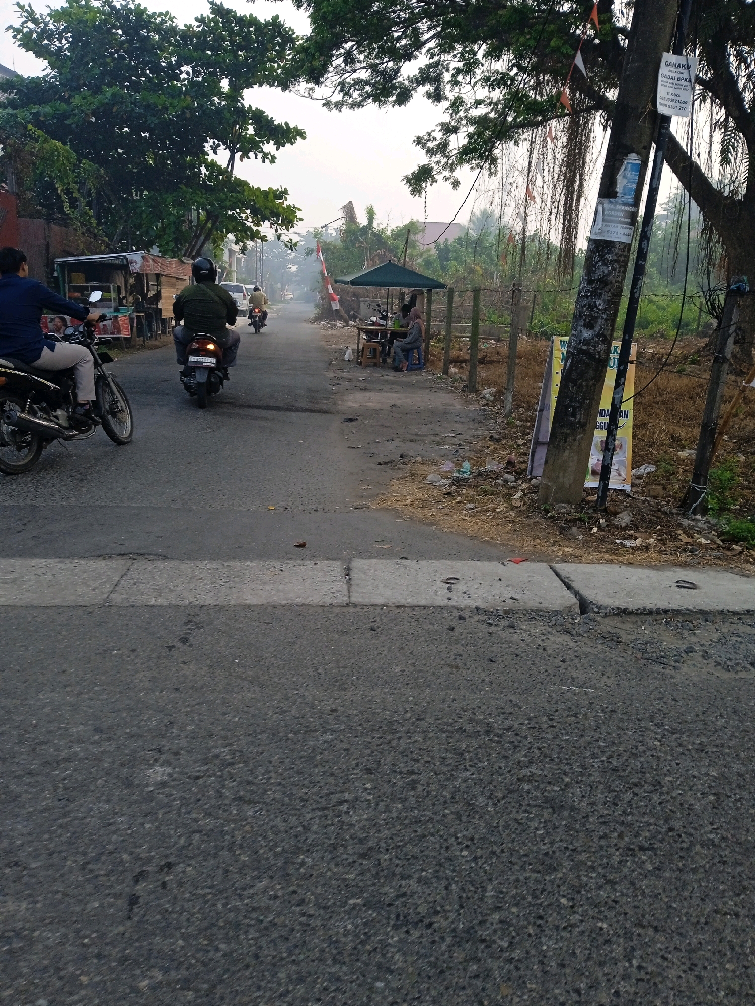 A street scene in Banjarmasin, Indonesia, captures the early morning haze. Two individuals on motorcycles are traveling down the road, with one in the foreground on the left, and another slightly further ahead in the center. To the right, a small open-air food stall is set up under a green canopy, with a few patrons seated at tables. Beyond the stall, a utility pole stands tall, adorned with various signs and banners. Trees line the sides of the road, with one prominent tree on the right exhibiting a dense canopy and hanging vines. The road itself appears to be paved and in fair condition, with a pedestrian crossing marked in the foreground. The overall atmosphere suggests a typical day in a bustling Indonesian city, with the presence of local commerce and everyday transportation.