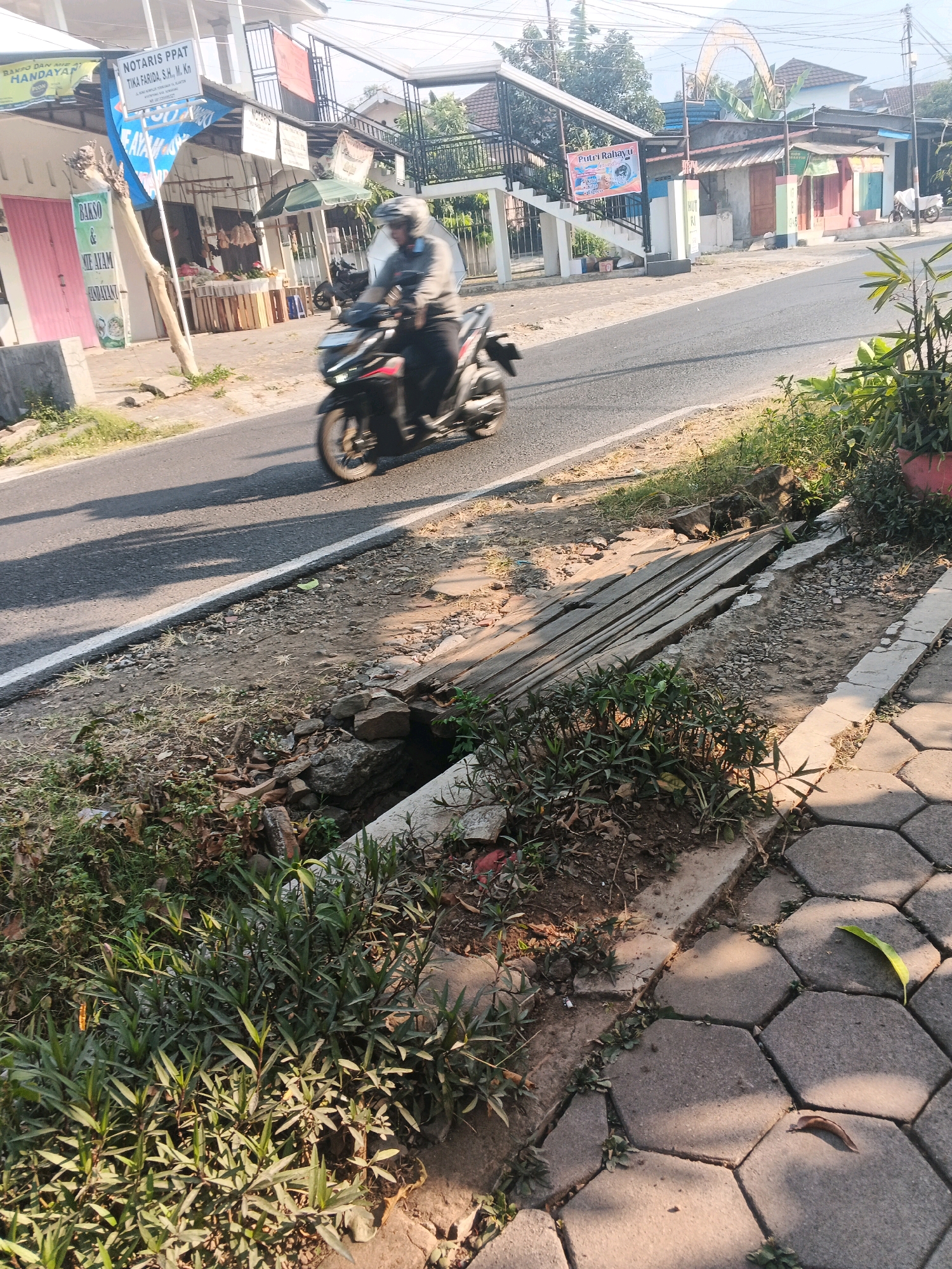 In Ungaran, Indonesia, a person wearing a helmet is riding a motorcycle on a paved road. The motorcycle is dark-colored and is in motion, casting a shadow on the road. To the left of the road, there is a small drainage ditch covered with wooden planks. Adjacent to the ditch is a sidewalk made of hexagonal pavers. Along the side of the road, there are various small shops and buildings. One building has a sign that reads "NOTARIS PPAT TIKA FARIDA, S.H., M. Kn." and another sign displays "BAKSO DAN MIE AYAM HANDAYANI." Further down the road, there are more buildings with colorful facades. The environment is a typical street scene in a town or city, with vegetation growing along the roadside and a sunny sky overhead.