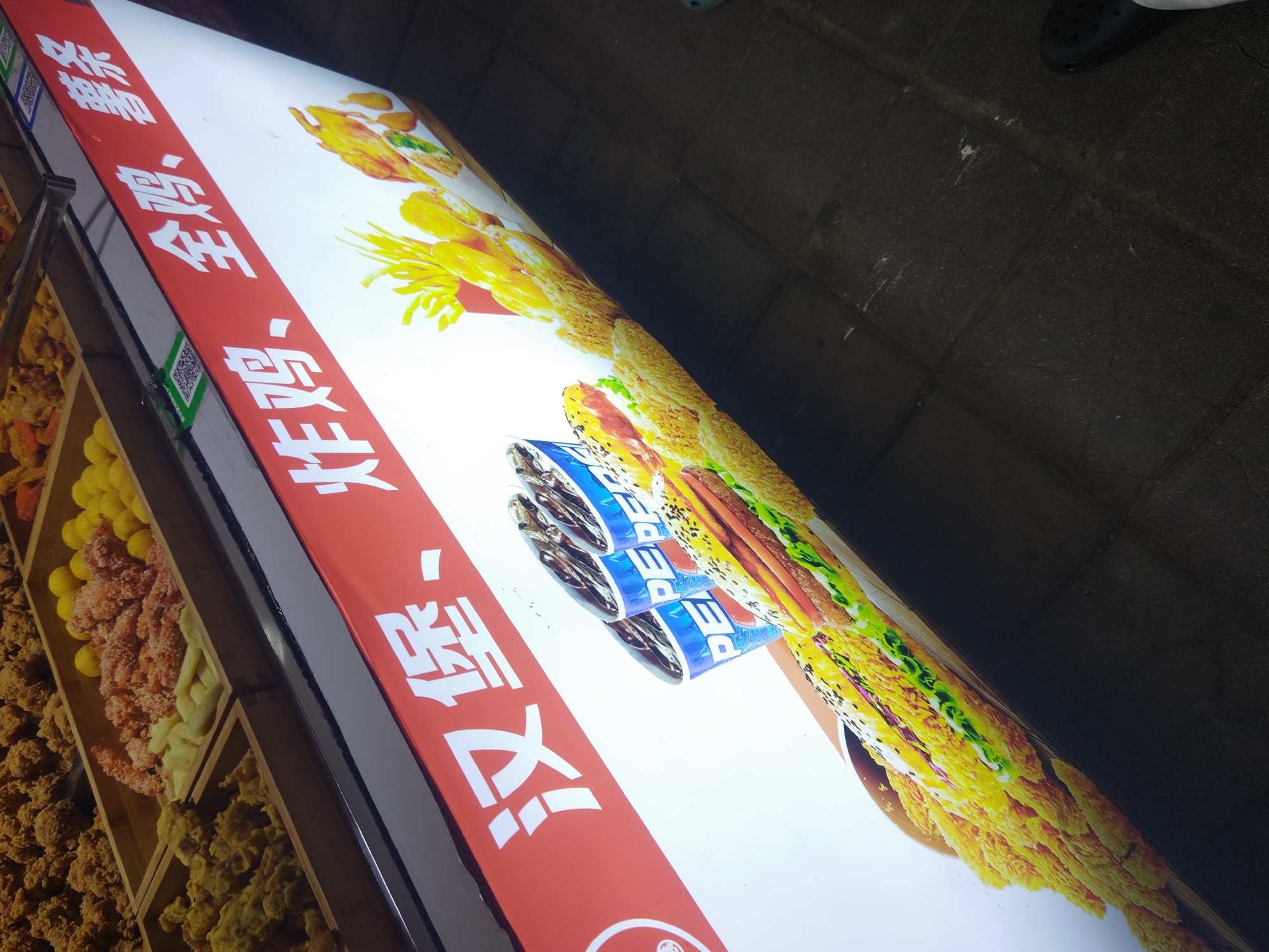 The image shows a food stall in Shapingba District, China. A display case on the left showcases various fried foods, including chicken, potatoes, and other snacks. Adjacent to the display case is a brightly lit sign. The sign features images of fried food, a stack of Pepsi cans, and Chinese characters written in white on a red background. The sign advertises "炸鸡" (fried chicken) and "实惠” (affordable). The background shows a dark grey-paved surface, with a foot clad in a dark shoe visible in the top right corner. The image appears to be taken at night.