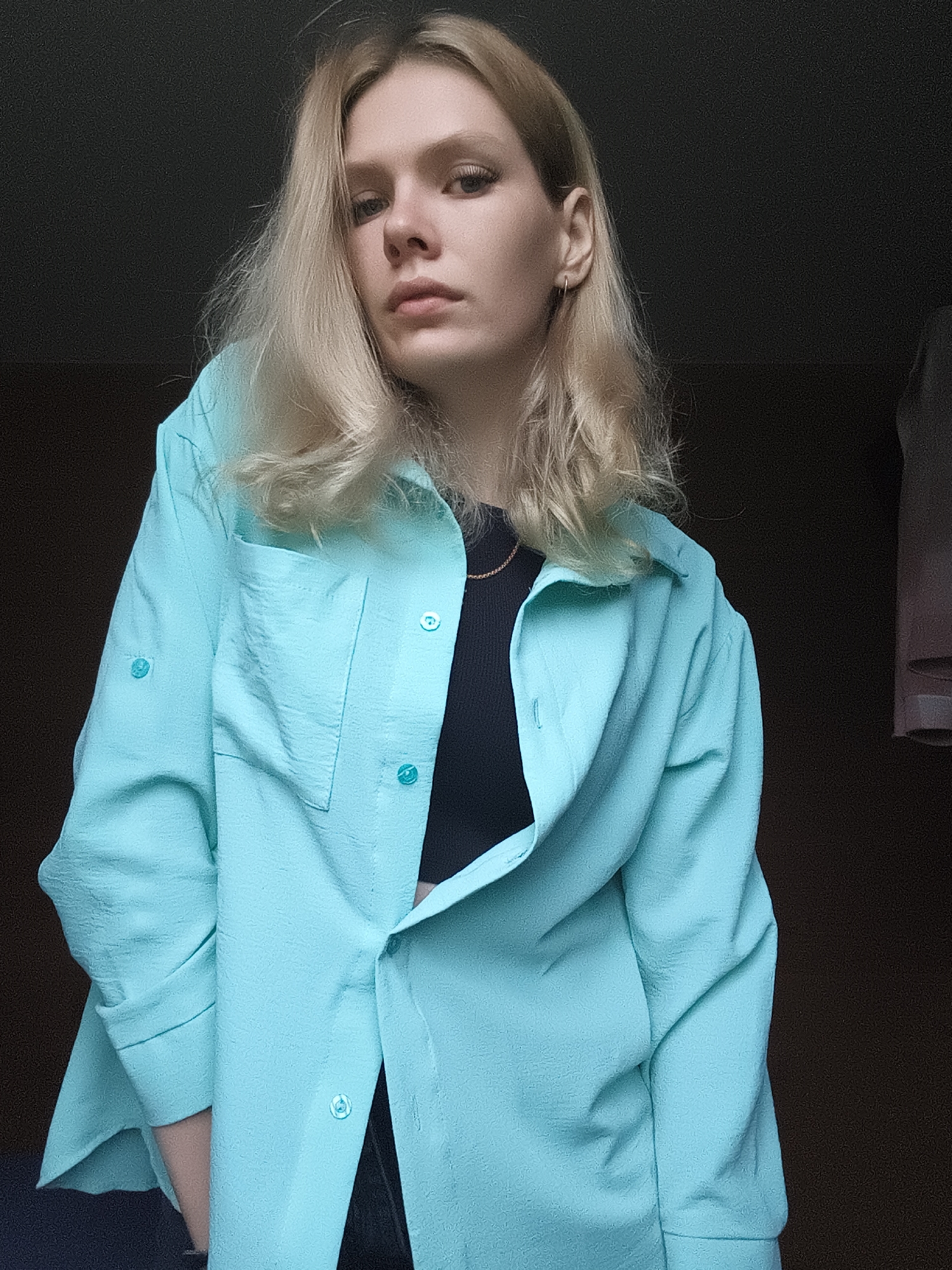 A young woman with blonde hair and light skin is posing for a photo indoors. She is wearing a bright turquoise button-up shirt over a black top. The shirt is open, revealing the black top underneath. She has minimal makeup on and is looking towards the camera. The background is dark and appears to be an indoor setting, possibly a room with a dark wall. There is some soft lighting on her face and upper body. The overall impression is a casual, candid portrait. The image does not contain any information that would allow for determining the specific location in Русский Вожой, Russia.