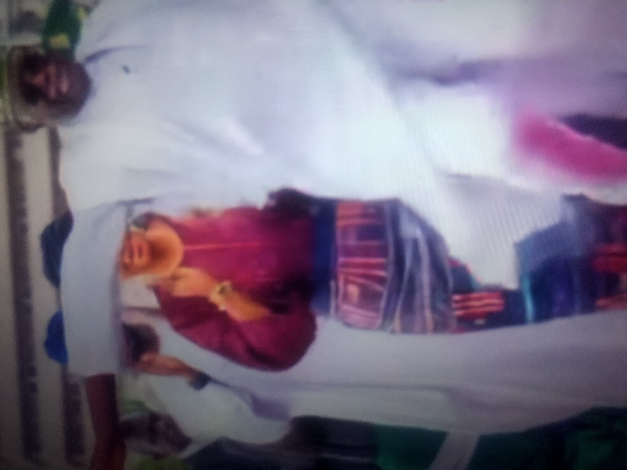 A low-resolution, color photograph shows a group of people in what appears to be a formal gathering, possibly outdoors. The primary subjects are a man in the foreground wearing a white flowing robe and a traditional cap, and a woman in the middle ground wearing a dark red top and a patterned skirt. The man in the foreground is looking to his right and appears to be smiling. The woman is also looking to her right, and her hand is raised as if gesturing or interacting with someone.
Several other individuals are partially visible in the background, also in white attire. The environment is indistinct, but there are hints of a building or structure with horizontal lines in the upper portion of the image. The overall impression is one of a public event or ceremony. No specific landmarks or features are discernible that would allow for precise identification of the location within Howayi, Nigeria. The lighting suggests it is daytime, but the resolution limits the ability to determine the specific time or atmospheric conditions.