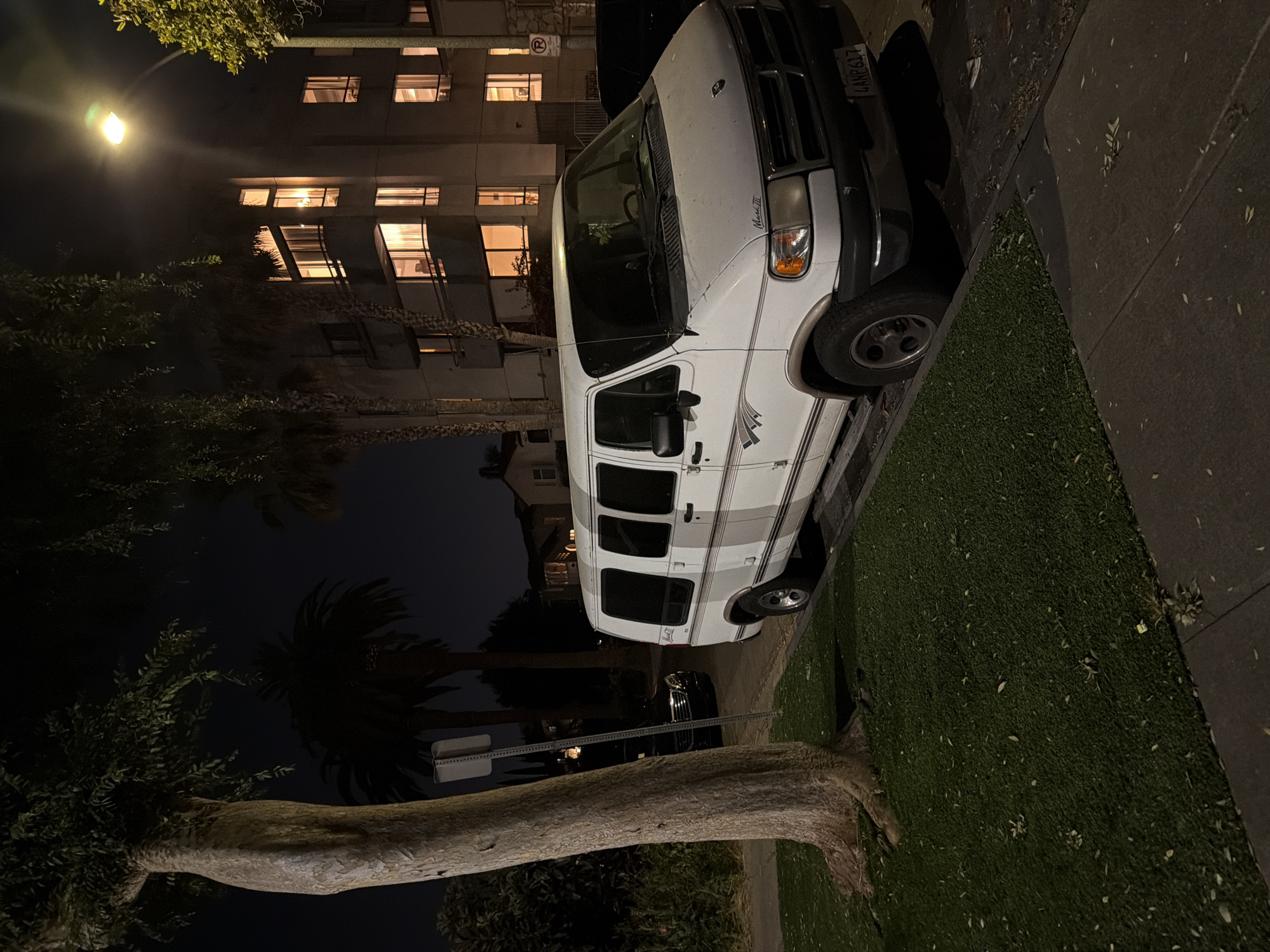 A white van is parked on a street at night, with the building and trees in the background. The van appears to be a cargo or passenger van, with tinted windows and striping along its side. The building behind the van has multiple windows, some of which are lit, suggesting it is occupied. The street is lined with trees, including palm trees, and there is a sidewalk and a patch of grass. A street lamp is visible in the upper left corner of the image, illuminating the scene. The overall impression is one of a quiet urban or suburban night.
There is text visible on the license plate of the van, which reads "4ANP617". The model name "Mark III" is visible on the side of the van. A circular sign with a red border and a white "R" in the center, likely a "No Parking" sign, is also visible. The image is taken at night, and the lighting suggests it is either dimly lit or illuminated by artificial light sources like street lamps and interior lights from the building. The weather appears to be clear.