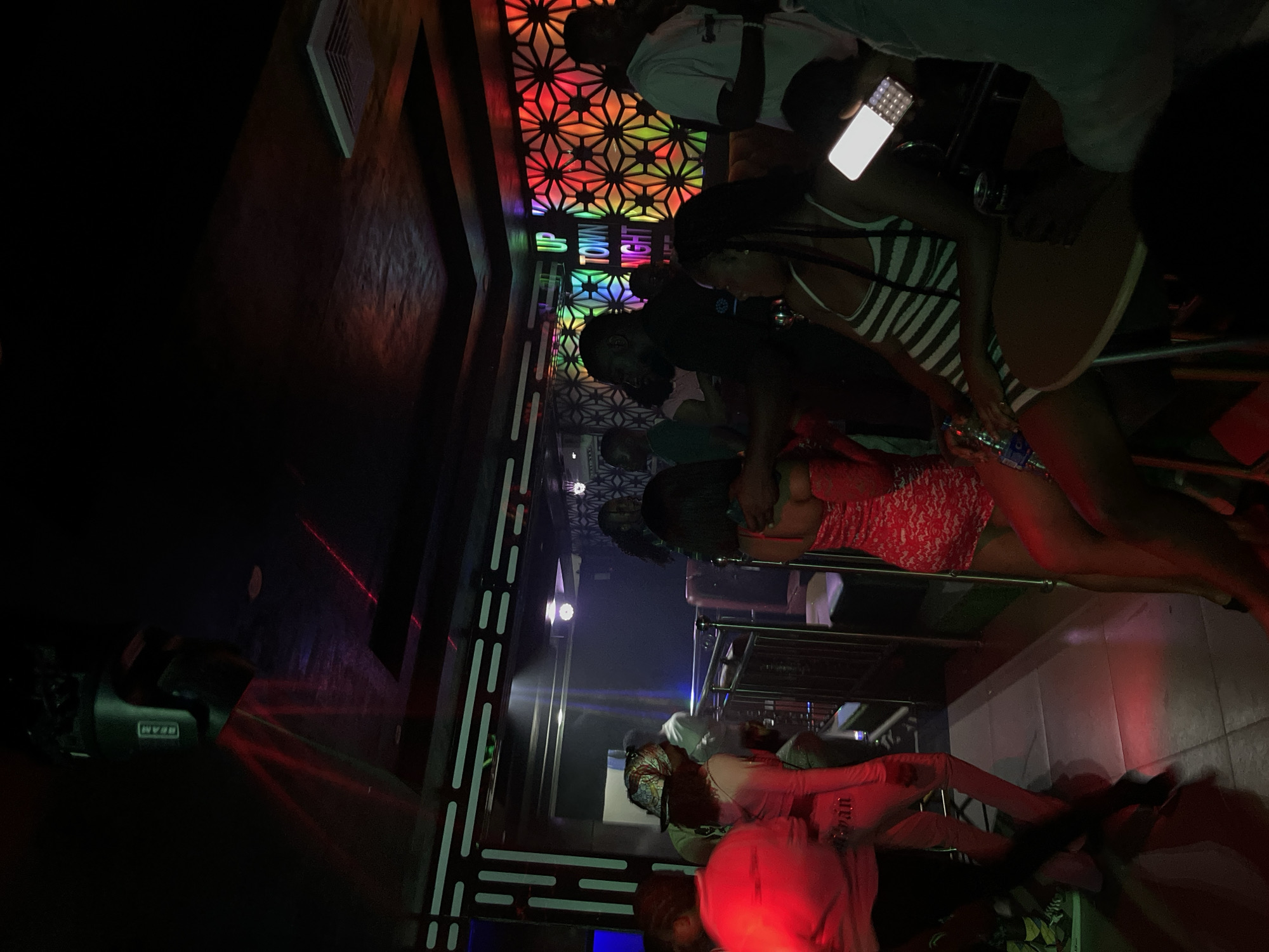 The image depicts a dimly lit nightclub environment with several individuals present. In the foreground, a person in a red lace dress is visible, and next to them, another person in a striped dress is holding a smartphone. To the left, a person in a bright red shirt is partially visible. The background features a colorful, geometric wall design and a neon sign that reads "UP TOWN NIGHT." Several people are standing and milling about, and the atmosphere suggests a social gathering or party. Lighting appears to be coming from overhead spotlights and the colorful wall.