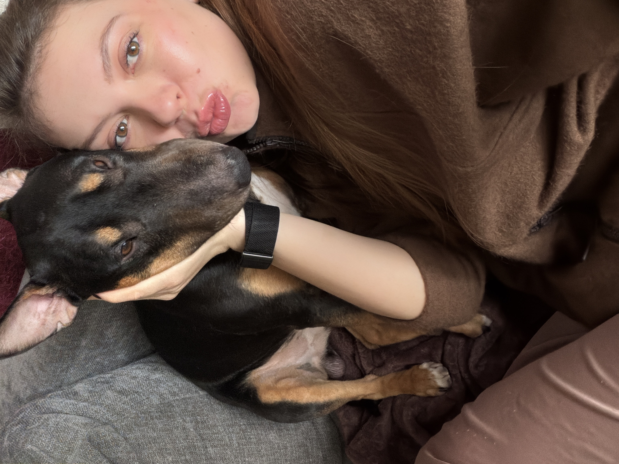 A close-up, high-angle shot captures a young woman and a dog in an intimate, cozy setting, likely indoors on a couch or bed. The woman, with light brown hair and a fair complexion, lies on her side, looking towards the camera with warm brown eyes. Her lips are puckered in a kissy face, and her left arm is wrapped around the dog. She wears a black digital watch on her left wrist and a soft, brown, possibly fuzzy, garment.
The dog, a small to medium-sized breed with short black and tan fur (resembling a Dachshund mix or similar terrier type), rests its head close to the woman's face and upper arm. Its dark eyes are partially visible, looking upwards. Its body is nestled against the woman, appearing relaxed and comfortable. The woman's hand gently cups the dog's head.
The background consists of textured fabrics: a grey, woven material directly beneath the dog and a section of the woman's brown garment or a brown blanket. The overall mood is one of affection and companionship. The lighting suggests an indoor setting, possibly during the day. No text is visible in the image. The location context provided is Khimki, Russia.