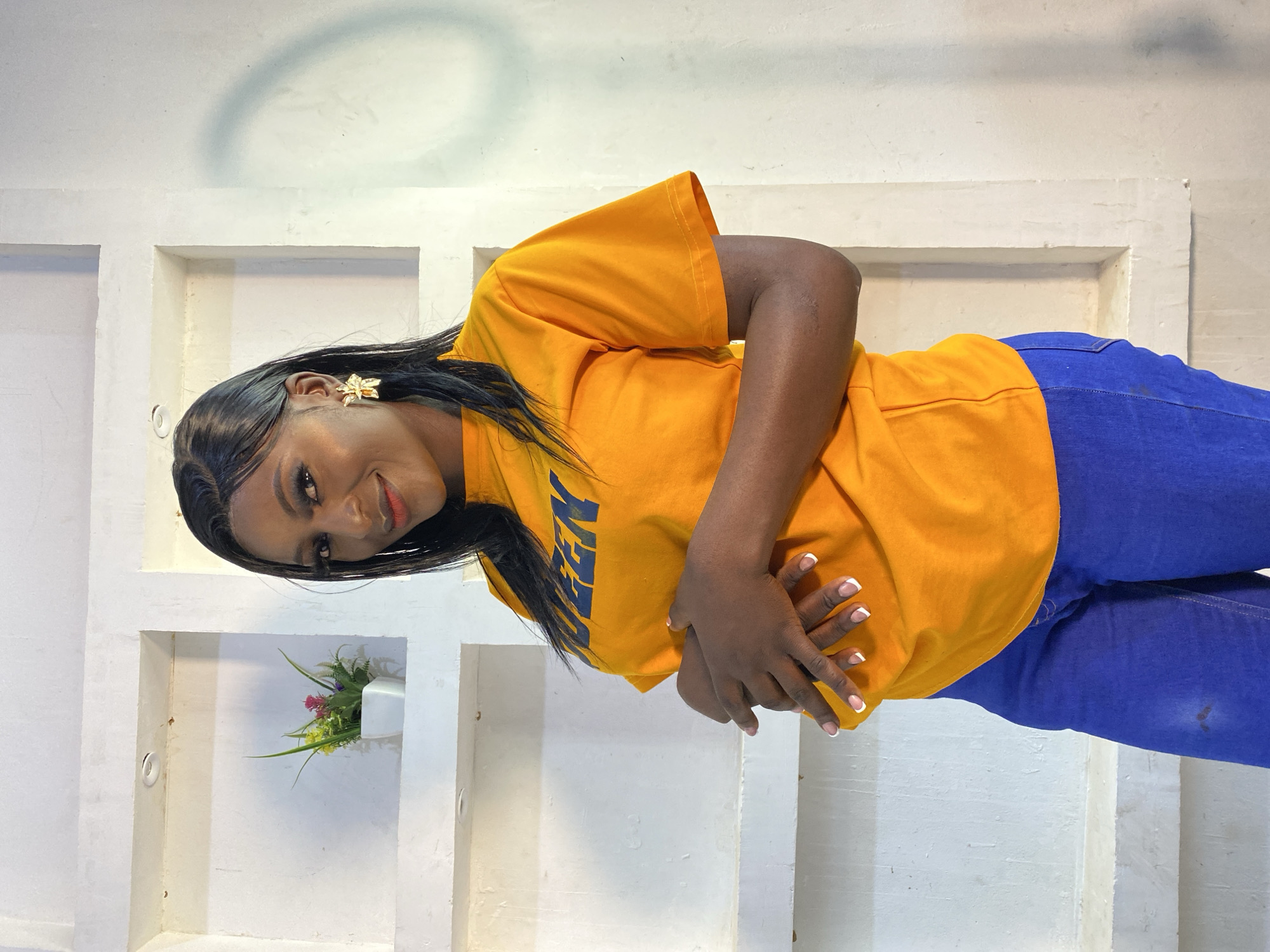 A medium shot shows a Black woman with long dark hair and dark skin posing in front of a white textured wall with recessed shelves. She is wearing an orange t-shirt with blue lettering that appears to spell "QUEEN", and royal blue jeans. Her hands are clasped over her stomach, and she is looking at the camera with a slight smile. She is wearing large, gold, floral-shaped earrings. A small potted plant with green and pink artificial flowers is visible in one of the wall's shelves. The lighting suggests an indoor setting, possibly a studio or residential space, with artificial light illuminating the subject and casting a shadow on the wall behind her. The environment does not contain any discernible landmarks or features that would place it specifically in Jos, Nigeria, or any other city.