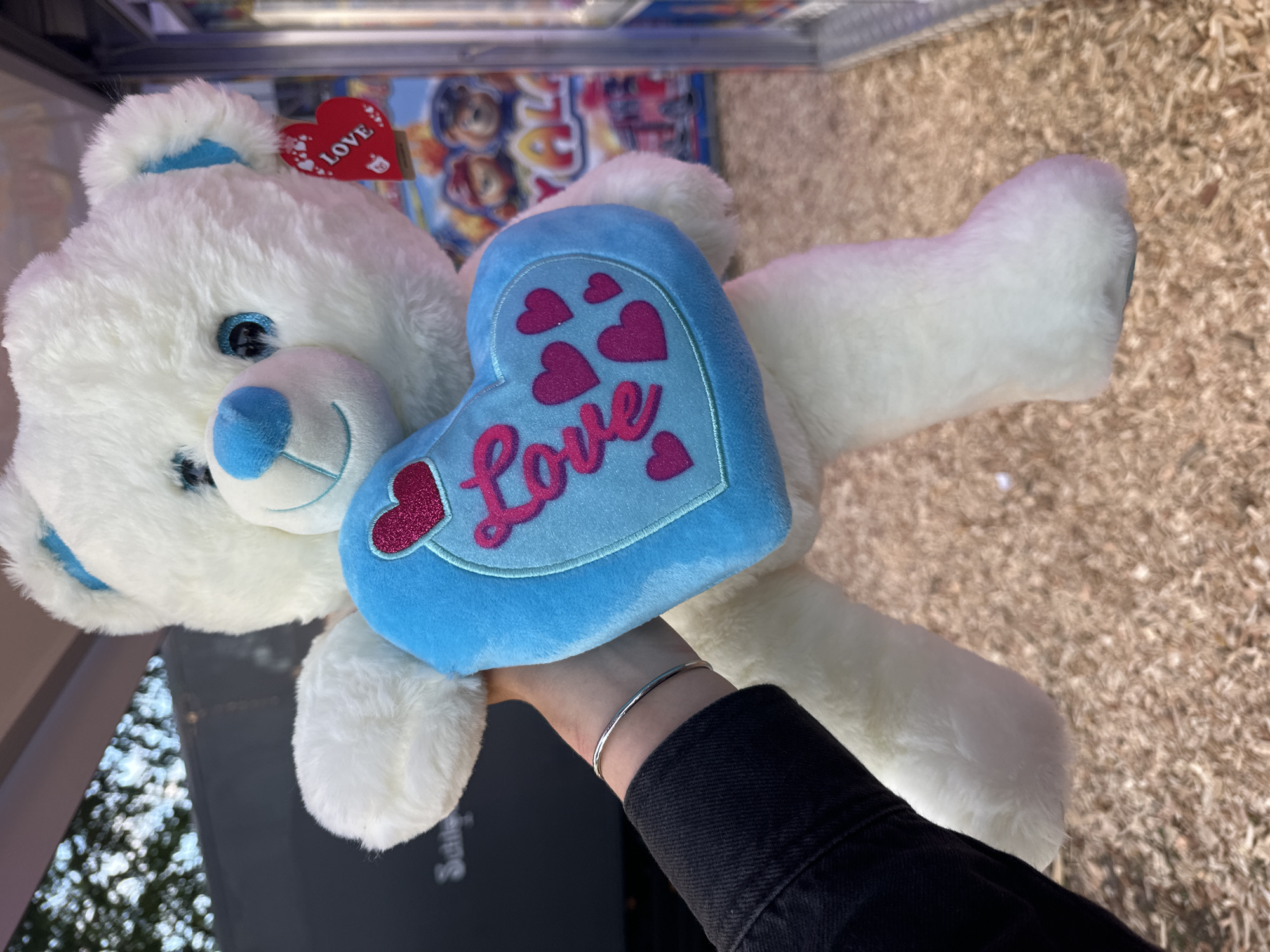 A white teddy bear with blue accents and a blue heart with "Love" written in pink cursive is held by a person's left hand, which is wearing a silver bracelet. The teddy bear is likely in an indoor setting, possibly a store or arcade, given the presence of a vending machine in the background. The ground appears to be covered in wood chips. The context of Bruchhausen-Vilsen, Germany, is not discernible from the image.