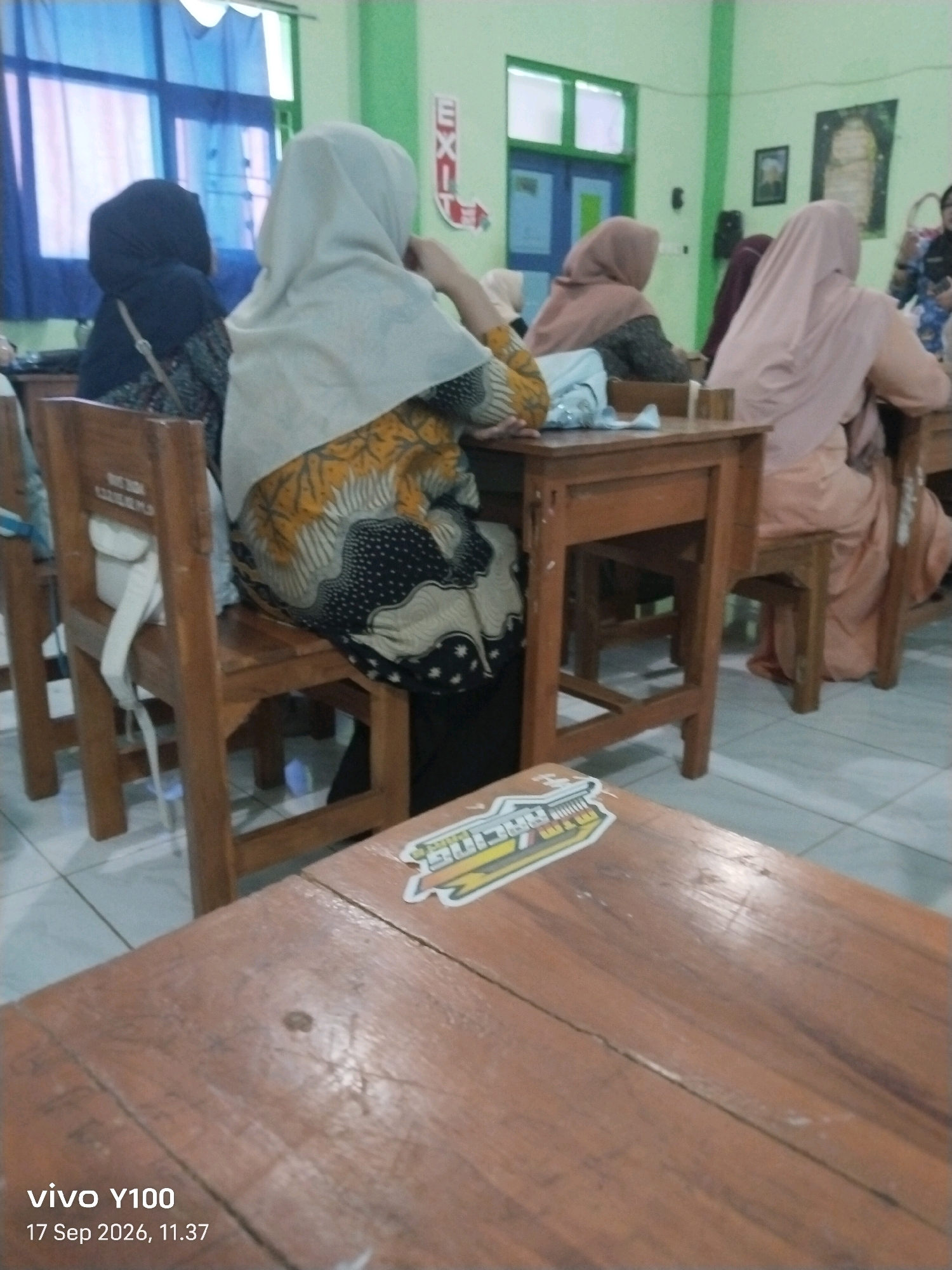 The image depicts a classroom scene in Situbondo, Indonesia, likely a school setting given the presence of desks and chairs. Several students, predominantly female, are seated at wooden desks, their backs to the camera. They are wearing headscarves, typical of the region. One student in the foreground wears a light-colored headscarf and a batik-patterned dress in shades of yellow, black, and white. Another student to her left wears a dark headscarf. In the background, other students are also visible, some in pastel-colored headscarves. The classroom walls are painted a light green, and a window with blue curtains is visible on the left. An "EXIT" sign is mounted on the wall. The floor is tiled. In the foreground, a wooden table has stickers on it, and the bottom of the image displays "vivo Y100 17 Sep 2026, 11.37".