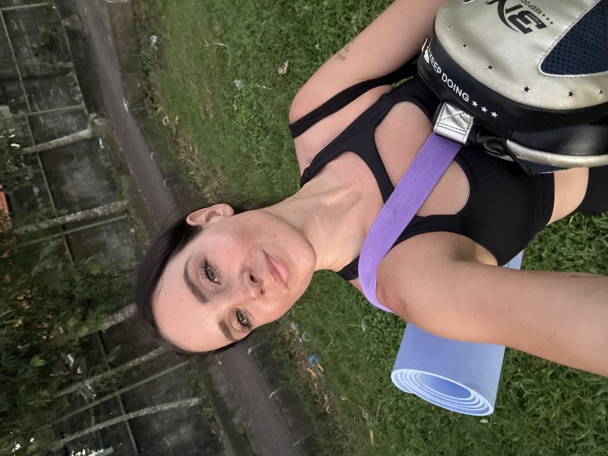 A woman with dark hair and light skin takes a selfie outdoors, in front of a low wall and trees. She is wearing a black sports bra and a purple strap across her chest. To her left, a rolled-up light blue yoga mat is visible. To her right, a black and silver boxing pad is slung over her shoulder. The ground is covered in green grass, and a paved path is visible behind her. The scene appears to be in a park or an outdoor recreational area in North Kuta, Indonesia.