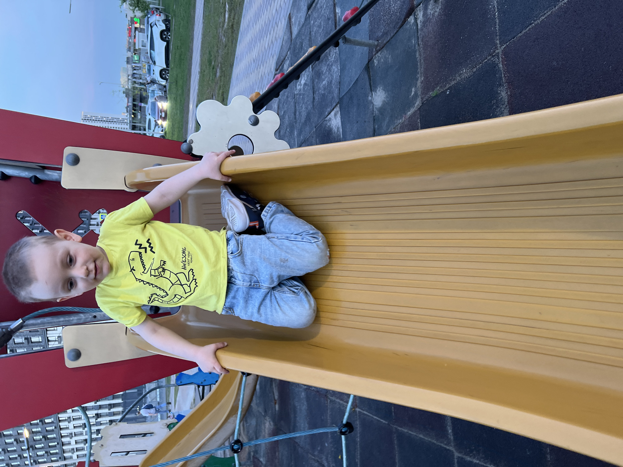 A child, positioned on a playground slide, is the central figure of the image. The child, a young boy, is wearing a yellow t-shirt with a dinosaur illustration and light blue jeans. He is holding onto the edges of the slide. The slide, made of yellow material, is the dominant object in the foreground.
In the background, a playground structure with red and beige components is visible, as well as sections of other playground elements like a climbing apparatus. Further in the distance, there are parked cars and apartment buildings, indicative of a city environment.
The image was taken at night, at the Children's playground at the intersection of 1st and 2nd Krugovaya streets in Mytishchi, Russia.
