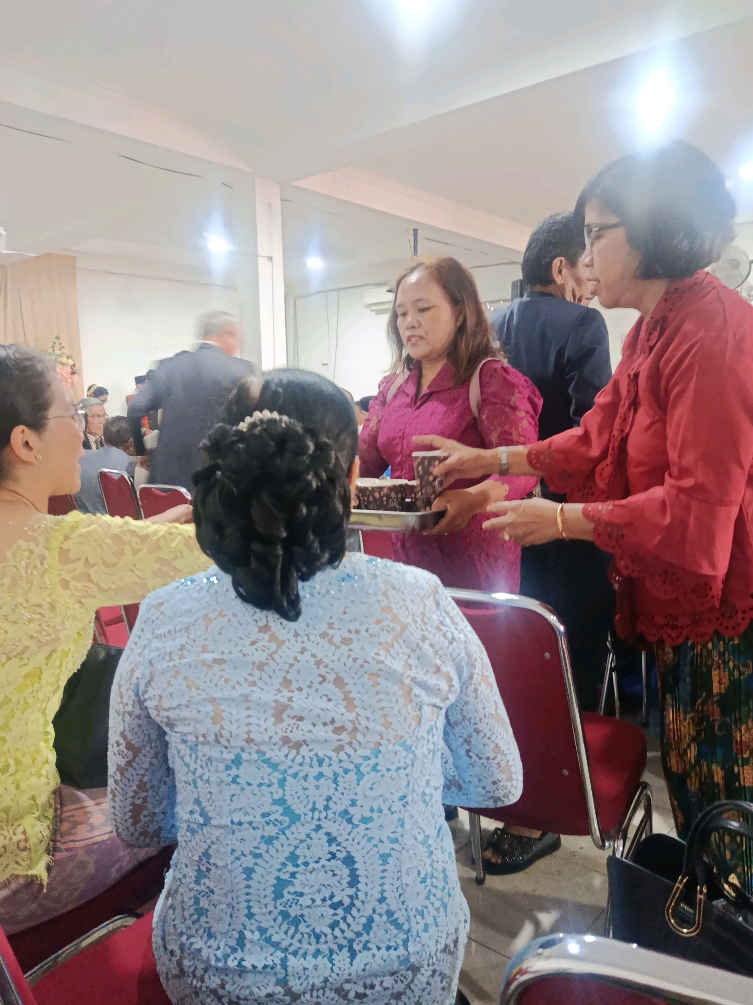 In Babelan, Indonesia, a social gathering is taking place indoors. The image focuses on several women dressed in traditional Indonesian attire, some of whom are seated and others standing. One woman in a bright yellow lace top is partially visible on the left, facing away from the camera. A woman in a light blue lace dress is seated in the foreground, her back to the camera, revealing an intricate lace pattern. To her right, a woman in a vibrant red top with decorative sleeves is interacting with another woman in a magenta outfit who is holding a tray of cupcakes. The environment appears to be a hall or reception area with red chairs and white walls, illuminated by bright overhead lighting. A man in a dark suit is blurred in the background, suggesting a larger event is in progress. A black handbag is visible in the lower right corner.