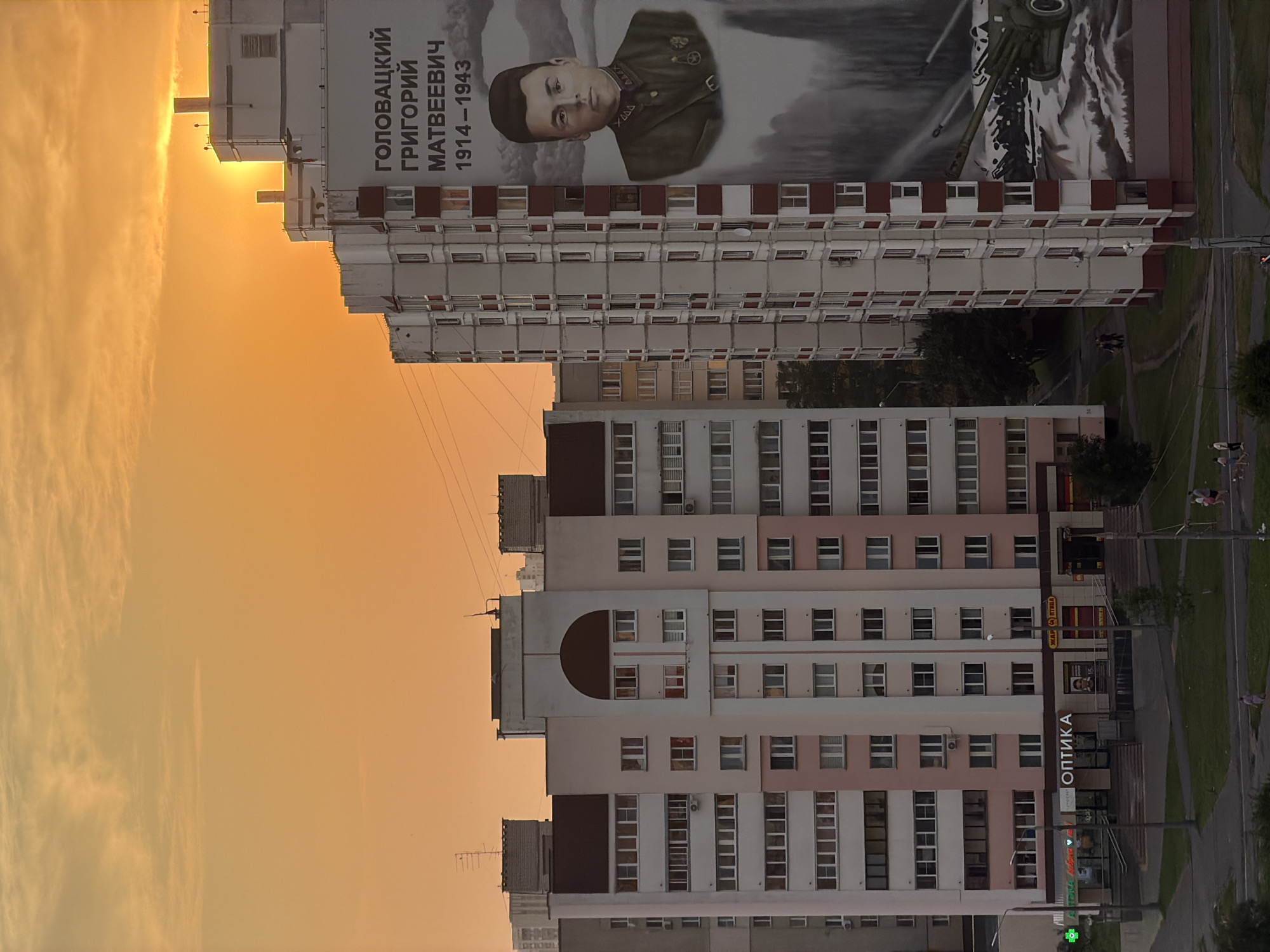 The image depicts a residential building in the city of Homyel, Belarus, at sunset. The sky is a vibrant orange, with the sun partially obscured by the top of the building. On the side of the building, a large mural features a portrait of a man, Grigory Matveevich Golovatsky, with his dates of birth and death (1914-1943). Below the portrait is an image of a military cannon. Other buildings, power lines, and streetlights are visible. In the foreground, there are grassy areas, a sidewalk, and a few people walking. A storefront with the sign "ОПТИКА" (Optika) is also visible at the base of one of the buildings.