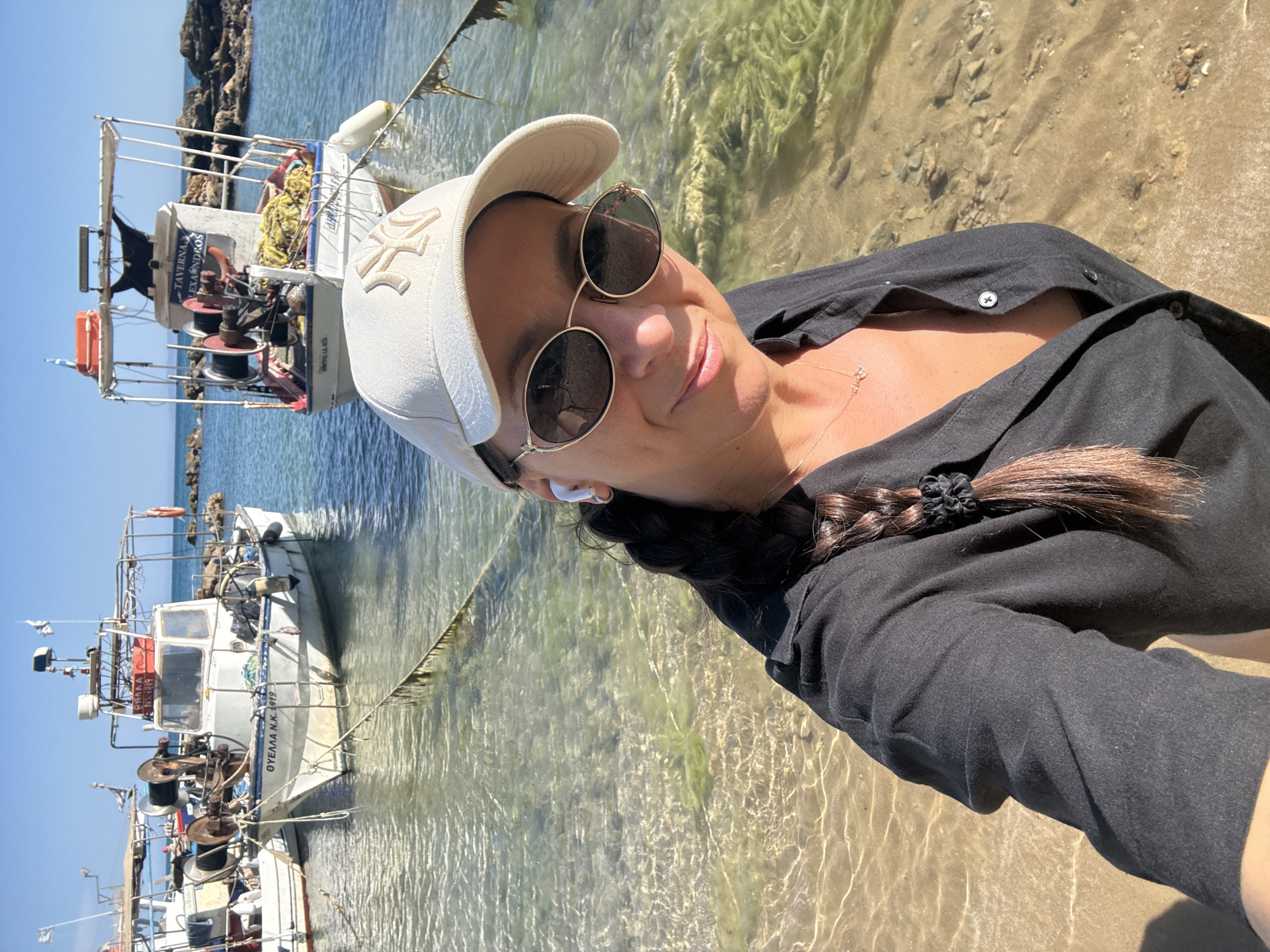 A woman with brown hair in a braid, wearing a black button-up shirt, sunglasses, and a light-colored baseball cap, takes a selfie on a sunny day at a beach. She is wearing a necklace and appears to be wearing wireless earbuds. Behind her, several fishing boats are docked in clear, shallow water. The environment is a coastline with sandy beach and rocky outcrops in the background. The fishing boats are white with various equipment on them, including nets and winches. One boat has the name "TAVERNA ALEXANDROS" visible on it, and another has "ΘΥΕΛΛΑ Ν.Κ. 1919" printed on its hull. The location is the Municipal Unit of Lefkimmi, Greece.