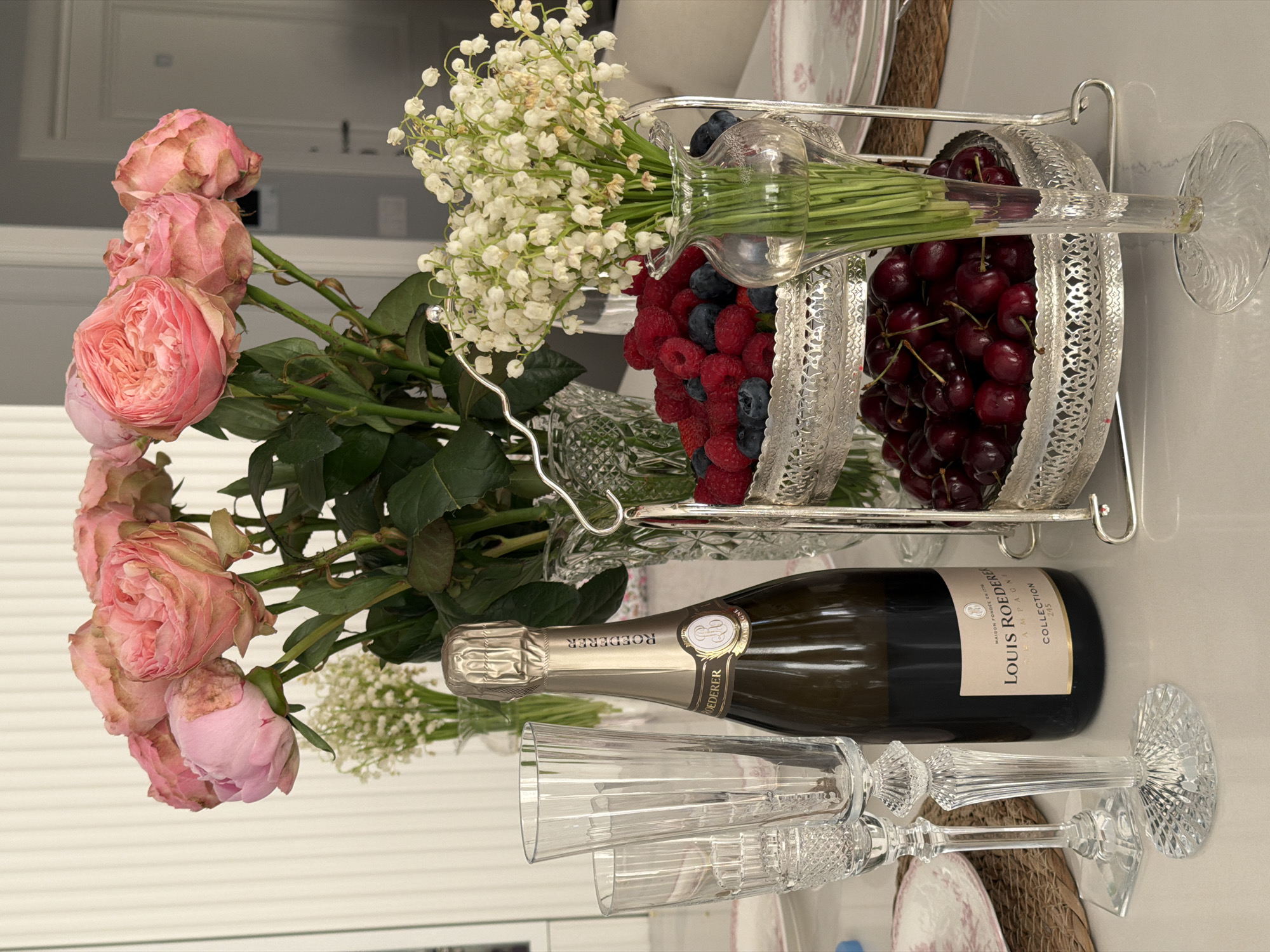 The image shows a table setting with flowers, berries, champagne, and glasses. A bouquet of pink roses and lily of the valley is on the left side of the frame. A silver tiered serving dish holds blueberries, raspberries, and cherries. A bottle of Louis Roederer Collection champagne lies horizontally, with two empty champagne glasses beside it. The setting suggests a celebration or special occasion, with attention to detail in the floral arrangement and drink selection. The background shows a glimpse of an interior, including a white door. There is no visual indication of Moscow or any location-specific details in the image.