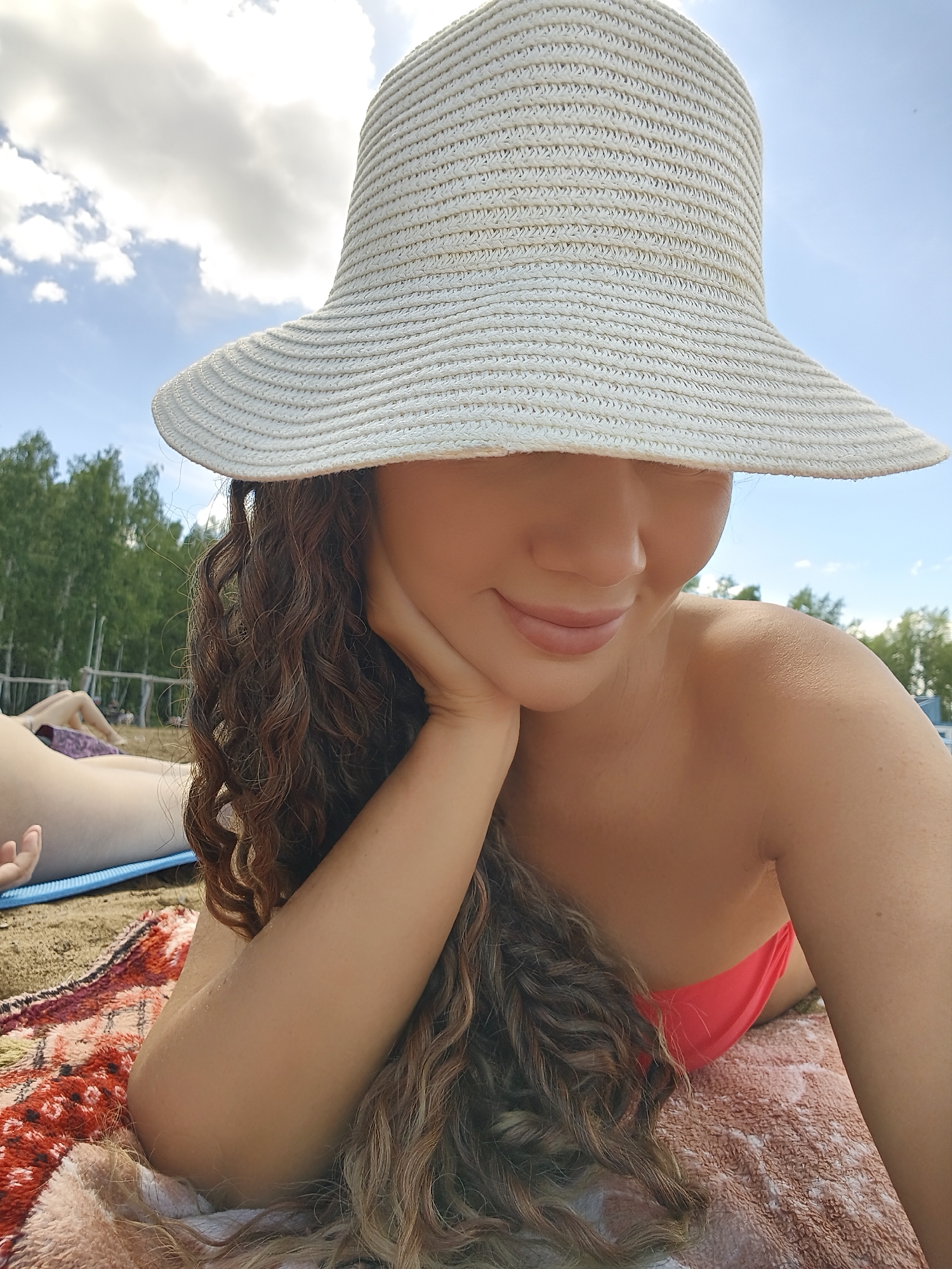 A young woman with long, curly brown hair lies on her side on a blanket at the beach. She is wearing a wide-brimmed straw hat that covers her eyes and a pink bikini top. Her right hand is propped under her chin, and she is looking down with a slight smile. Another person is partially visible in the background, lying on a blue mat. The scene appears to be a sunny day, with a bright blue sky and fluffy white clouds. Trees are visible in the background, suggesting a wooded area near the beach. The overall impression is one of relaxation and enjoyment of a summer day.