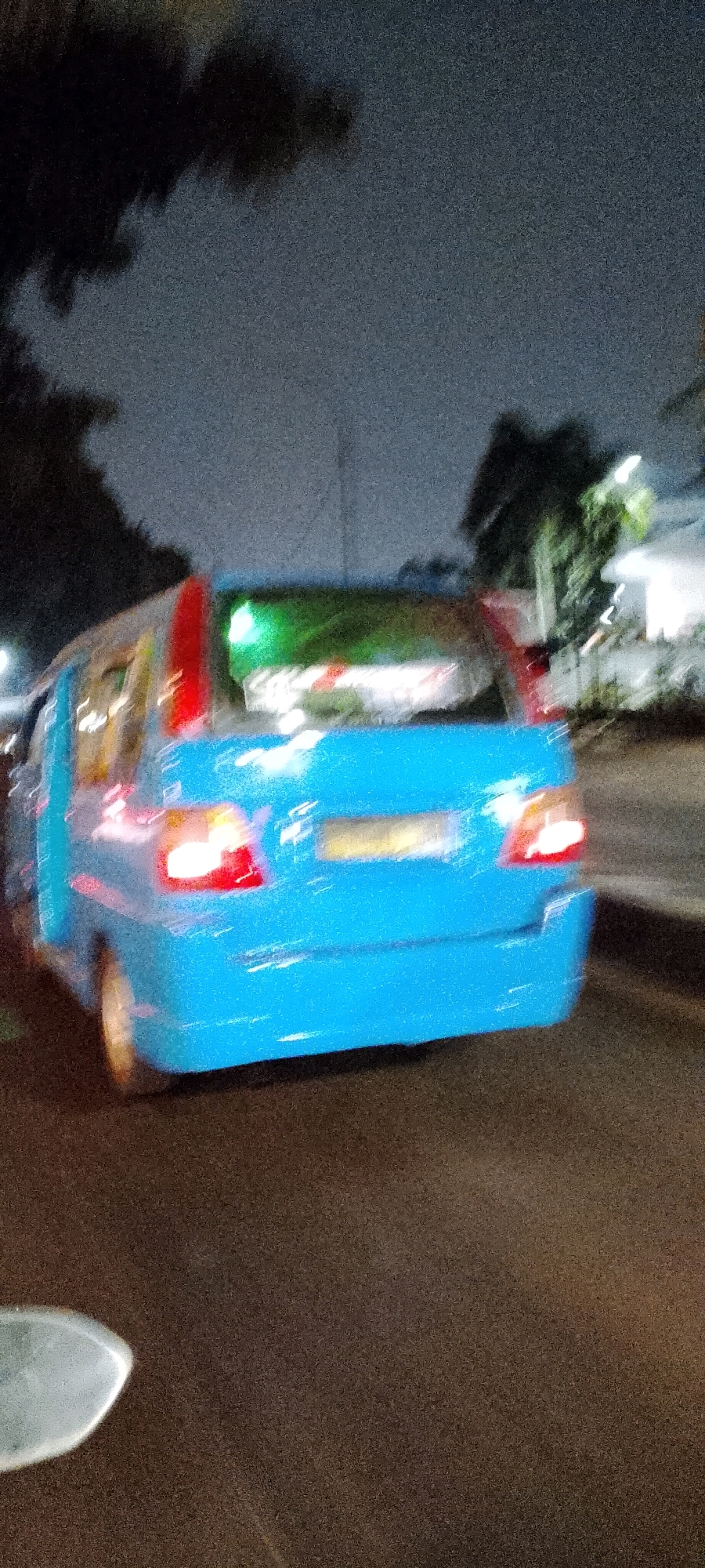 A bright blue van is captured in motion on a city street at night. The van has red accents on its roof and taillights, and a green light is visible on the roof above the rear windshield. A yellow license plate is present on the back of the vehicle. The scene is blurry, indicating movement and potentially low light conditions. Trees and some street lights are visible in the background. The overall setting appears to be an urban environment in Depok, Indonesia.