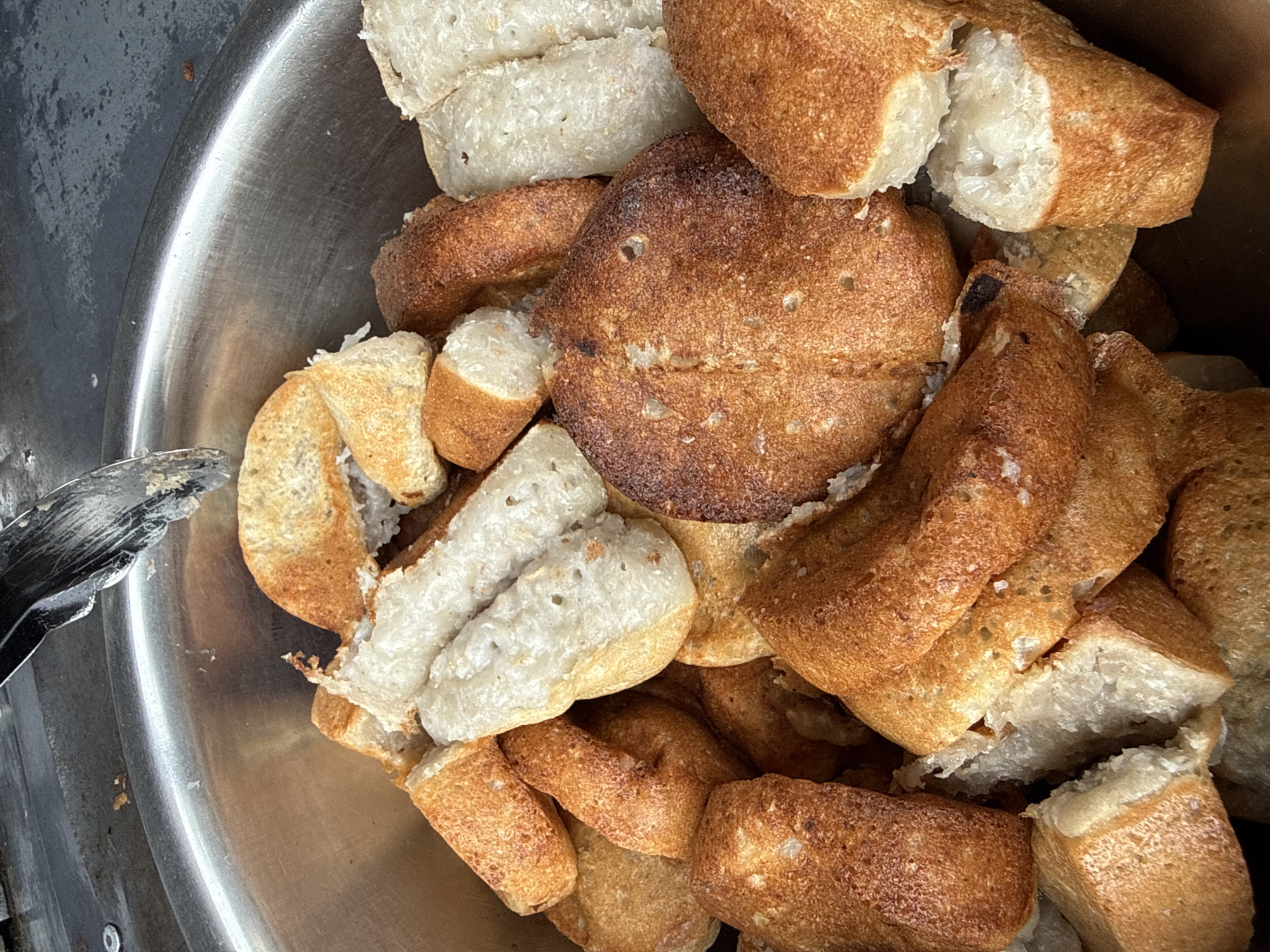 A close-up, overhead view shows a large, stainless steel bowl overflowing with freshly baked kuih bangkit, a traditional Indonesian cookie. The kuih bangkit are varied in shape and size, with some being round and flat, while others are elongated and cylinder-like. They exhibit a golden-brown exterior, indicating they have been baked to perfection, and a white, airy interior. The texture appears slightly crisp on the outside and soft within. The bowl is situated on a flat, dark surface, possibly a tabletop or counter, with a metal tong visible at the top left corner, suggesting recent handling. The overall impression is one of abundance and deliciousness, hinting at a market stall or a home kitchen preparing for a celebration. Without additional visual cues, the specific location within Lhokseumawe, Indonesia, cannot be determined from the image alone.