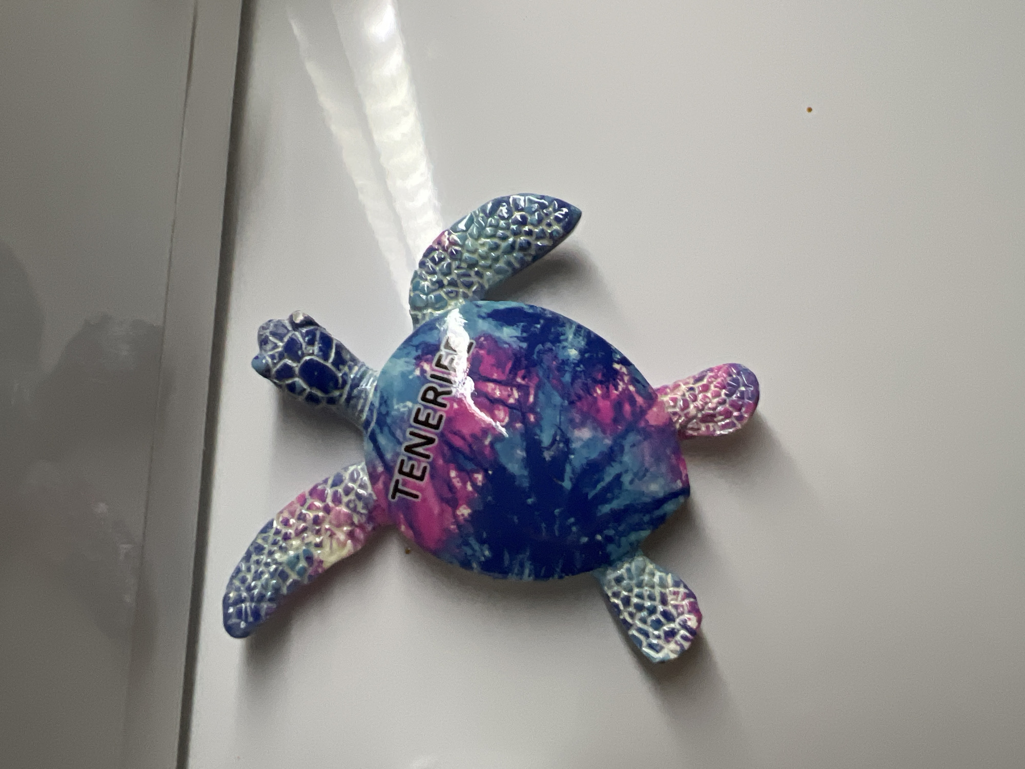 The image features a colorful, turtle-shaped decorative item, likely a magnet, placed against a plain, light-colored vertical surface. The turtle is adorned with a vibrant, abstract pattern in shades of deep blue, magenta, and purple, resembling a tie-dye or watercolor effect on its shell. Its four flippers display a distinct, raised, scale-like texture, also colored with similar blue, pink, and off-white tones.
Vertically written in white capital letters across the turtle's shell is the word "TENERIFE," indicating its origin or theme as a souvenir from the Canary Islands. The turtle is oriented diagonally, with its head pointing towards the upper left of the frame.
The background surface is smooth and appears to be a light grey or off-white, possibly a refrigerator door or a magnetic board, and exhibits subtle reflections or glare on the left side, suggesting indoor lighting. A faint vertical line runs along the left edge of the background. In the upper right quadrant of the background, a small, light brown dot is visible. There are no people present, and no activity is taking place; the image is a static display of the object.
