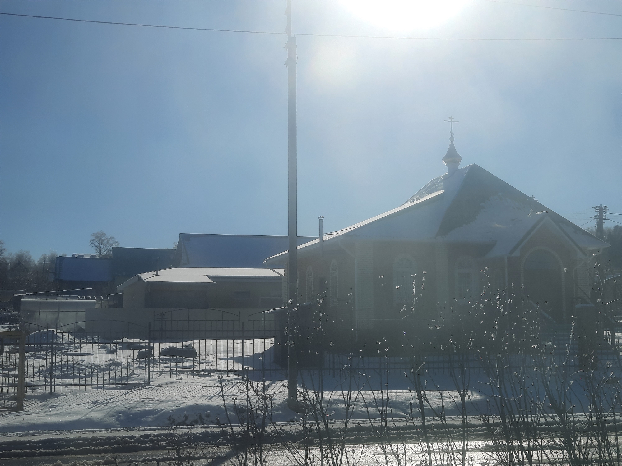 The image depicts a winter scene in Krasnogorskoye, Russia. A single-story wooden church with a dome topped by a cross stands prominently. The church and surrounding buildings have snow on the roofs. A black metal fence encloses the area in front of the church, and snow covers the ground. Bare bushes are in the foreground, partially obscuring the view. A utility pole rises on the left side of the church. In the background, other buildings are visible, also covered in snow. The sky is a clear blue with bright sunlight.