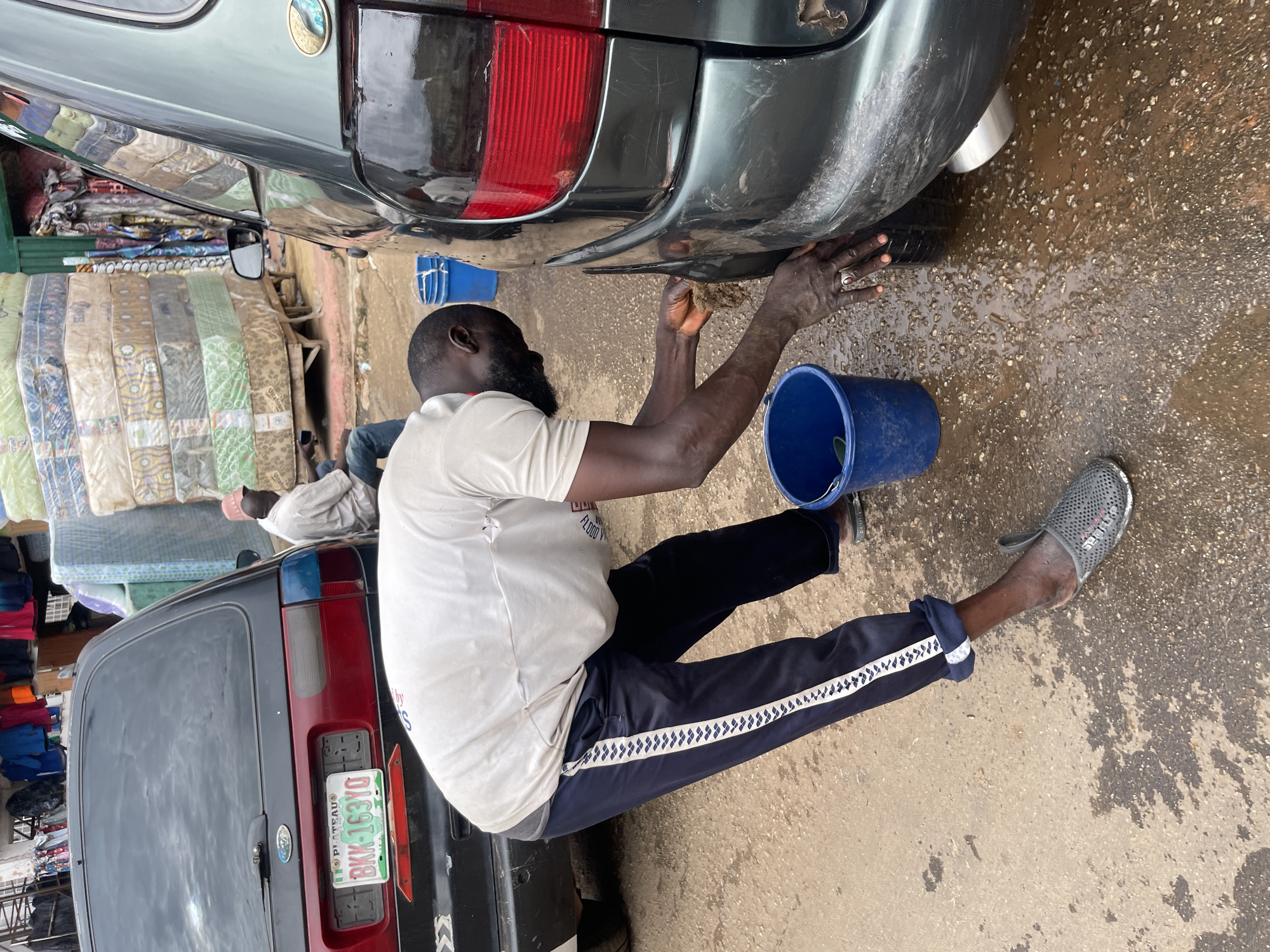 In Jos, Nigeria, a man is seen washing a car. He is wearing a light-colored t-shirt, dark blue track pants with a white stripe, and gray clogs. He is bent over, using his hands to scrub a section of the car's rear bumper. A blue bucket sits on the wet ground next to him, containing what appears to be soapy water. The car, a dark color, is parked on a paved surface that is wet and appears to be a road or parking area. In the background, stacks of mattresses are visible, suggesting a nearby market or storage area. Another person in a light-colored shirt and a pink hat is sitting on the ground in the middle distance. The overall environment suggests an outdoor, commercial setting in the city.