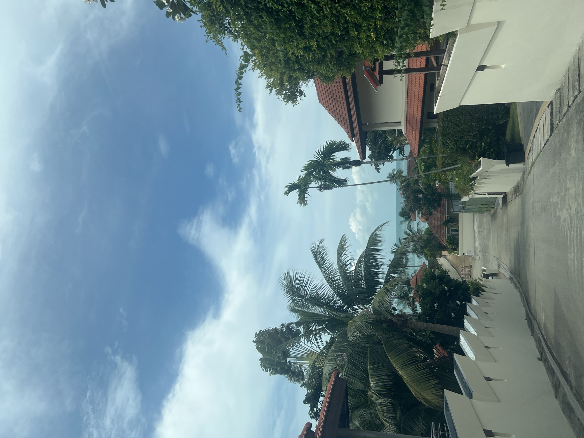 The image depicts a street winding downhill towards the sea, likely in the Ko Samui District of Thailand. The architecture suggests a resort or residential area, with several buildings featuring terracotta tiled roofs and white stucco walls. Lush tropical vegetation, including palm trees, is abundant, framing the view of the turquoise ocean in the distance. The sky is a mix of blue and white clouds, indicating pleasant weather. The street itself is paved and appears to be well-maintained. There are no people visible in the scene.