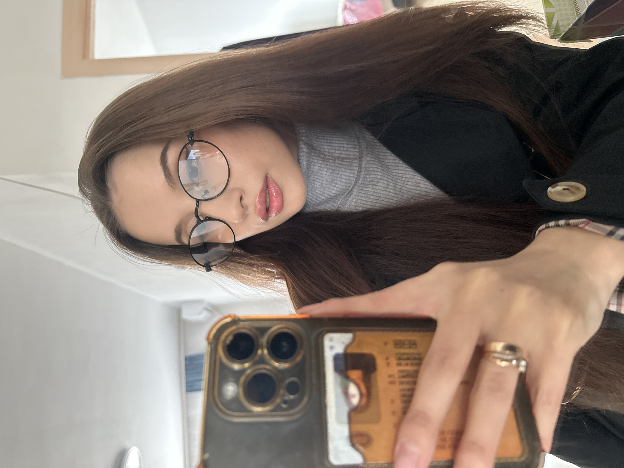 A young woman with long brown hair and glasses is taking a selfie. She is wearing a gray turtleneck sweater and a black coat. She is holding an iPhone with a gold and gray case. She is wearing rings on her finger. The background is blurry and shows a white wall with a mirror. The location is likely indoors and not identifiable as Yekaterinburg.