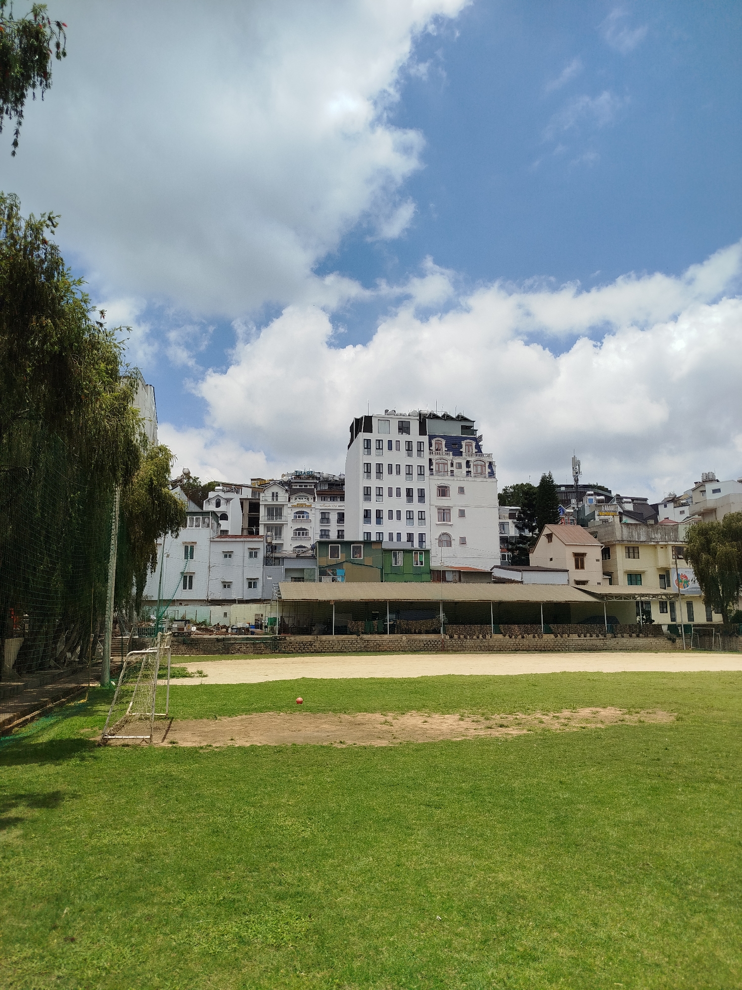 The image shows a grassy field with a soccer goal on the left. An orange soccer ball rests on the field closer to the middle. Behind the field are buildings of various heights, mostly white, in Da Lat, Vietnam. A row of covered seating is present at the edge of the field. The sky is blue with white clouds. A tree is on the left edge of the frame.