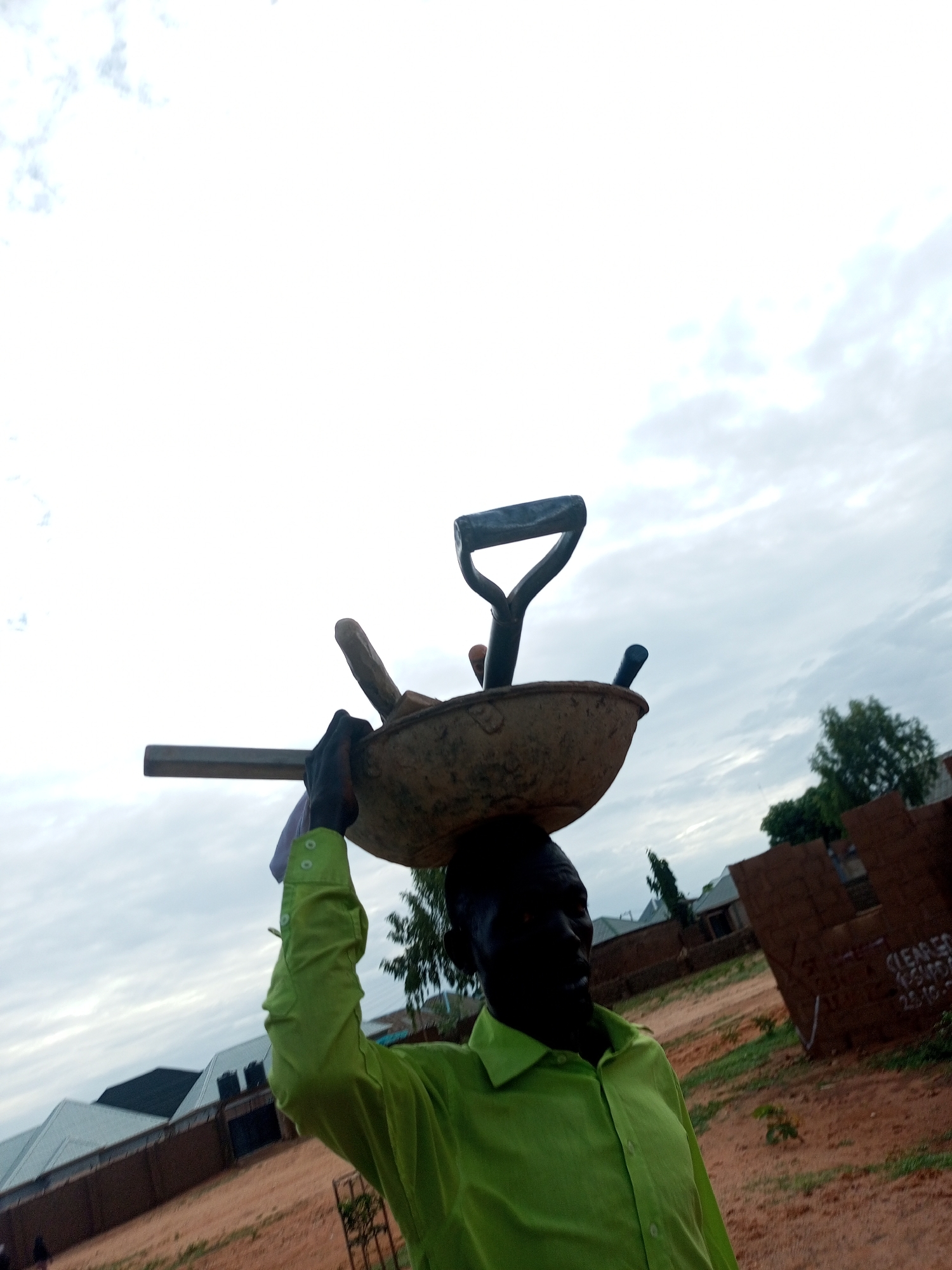 The image depicts a single individual, likely an adult male, of dark complexion, oriented slightly to the right of the frame's center. The person is wearing a long-sleeved, collared, lime-green shirt and is carrying a load on their head, supported by their raised left arm. The load consists of a dirty, light-brown, shallow metal basin (possibly a head pan) containing multiple tools. A "D"-grip shovel handle is visible, protruding upwards from the pan. Other tools include a wooden-handled implement and a smaller blue-handled tool. A long, rectangular object, possibly a metal bar, extends horizontally to the left from the pan, also supported by the person's left hand, which is wrapped in a light-colored cloth.
The scene is outdoors under an overcast sky with light grey and white clouds. The ground is reddish-brown earth with sparse green vegetation. In the background, to the left, there is a low wall made of brown blocks, behind which several buildings with grey or dark roofs and water tanks are visible, suggesting a residential or developing area. To the right, a large, dark-brown structure, possibly an unfinished wall or a pile of building materials, bears partially visible white text, including "CLEARED" and a date "23.10.2". Trees and bushes with green foliage are also present in the background. The location is in the city of Unknown, Unknown.