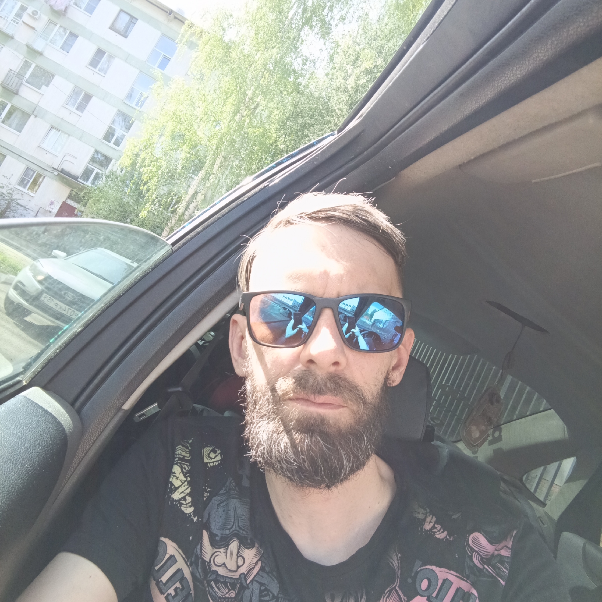 A selfie taken inside a car, likely in Chaikovsky, Russia, features a middle-aged man with a beard and sunglasses. He is wearing a black t-shirt with a graphic design. The reflection in his sunglasses suggests he is holding a phone. Outside the car window, there is a white apartment building with green trim and trees. Another car is also visible outside the driver's side window. The overall lighting suggests it is daytime.