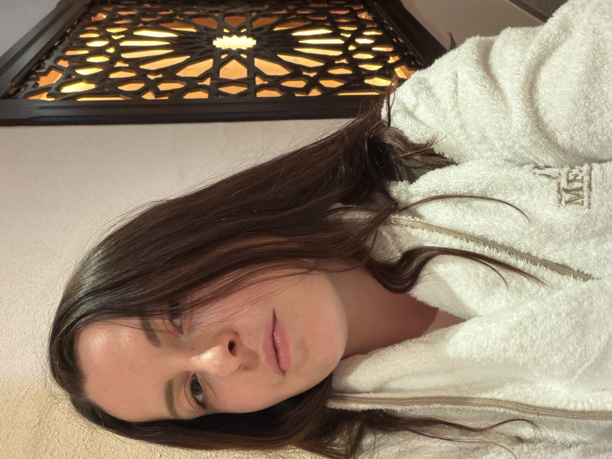 A person with long dark hair and light skin is wearing a white, fluffy robe with a brown zipper. The person is looking directly at the camera. The background includes a decorative ceiling panel with a geometric pattern. The location is Bergisch Gladbach, Germany.