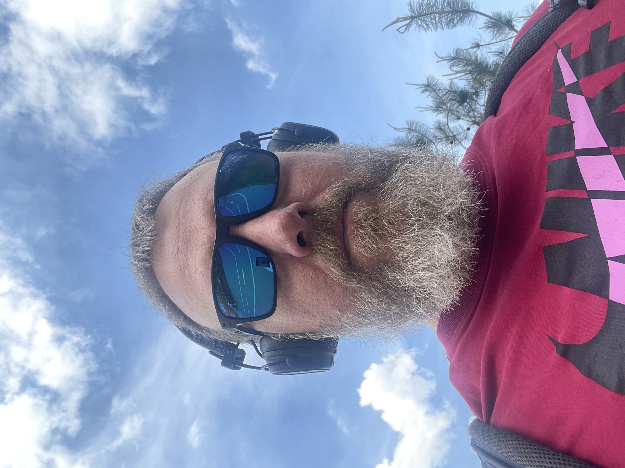 A low-angle selfie shows a fair-skinned man with a gray beard and gray hair wearing sunglasses and headphones. He is wearing a red long-sleeved shirt with a large, stylized, two-toned pink and black design on the right side. Above and behind the man, the bright blue sky is filled with fluffy white clouds. A few green pine needles are visible at the top right of the frame. The exact location of the city of Khimki, Russia is not evident in the photo.