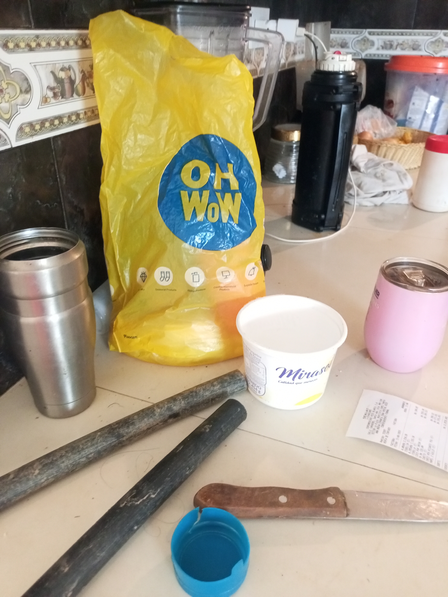 The image depicts a collection of household items on a tiled countertop. Prominently featured is a large, crinkled yellow plastic bag with a blue circular logo that reads "OH WoW". To the left of the bag, a brushed stainless steel tumbler is visible. In front of the bag is a white plastic tub with the brand "Mirasol" printed on it. To the right of the tub, a pink insulated tumbler is resting on a piece of paper, which appears to be a receipt. Behind the yellow bag, a black thermos and a blender are partially visible, along with a woven basket containing some items. In the foreground, two dark-colored cylindrical objects, possibly metal rods or pipes, lie diagonally across the counter. A bright blue plastic cap is also present near the bottom center of the image, and a knife with a wooden handle and a metal blade is positioned horizontally across the bottom right. The background shows a tiled wall with decorative elements. The scene appears to be an indoor setting, likely a kitchen or utility area. The presence of everyday objects suggests a domestic environment. The image does not contain any explicit indicators of the city of La Tagua, Venezuela, nor does it depict any people or specific actions.