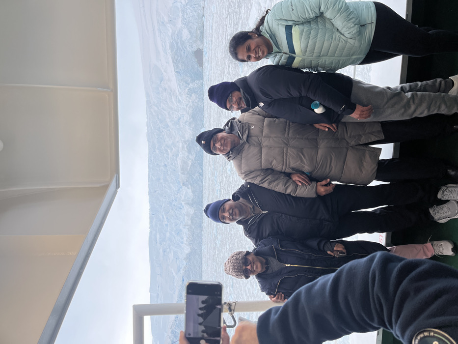A group of six people are standing near the edge of a boat, posing for a photo. They are dressed in winter attire, including jackets and hats, suggesting a cold climate. Behind them is a large, snow-covered expanse, possibly a glacier or icy landscape. One person is holding a smartphone, taking a photo of the group with the icy background in the frame. The scene suggests they are on a tour or expedition in a polar region.