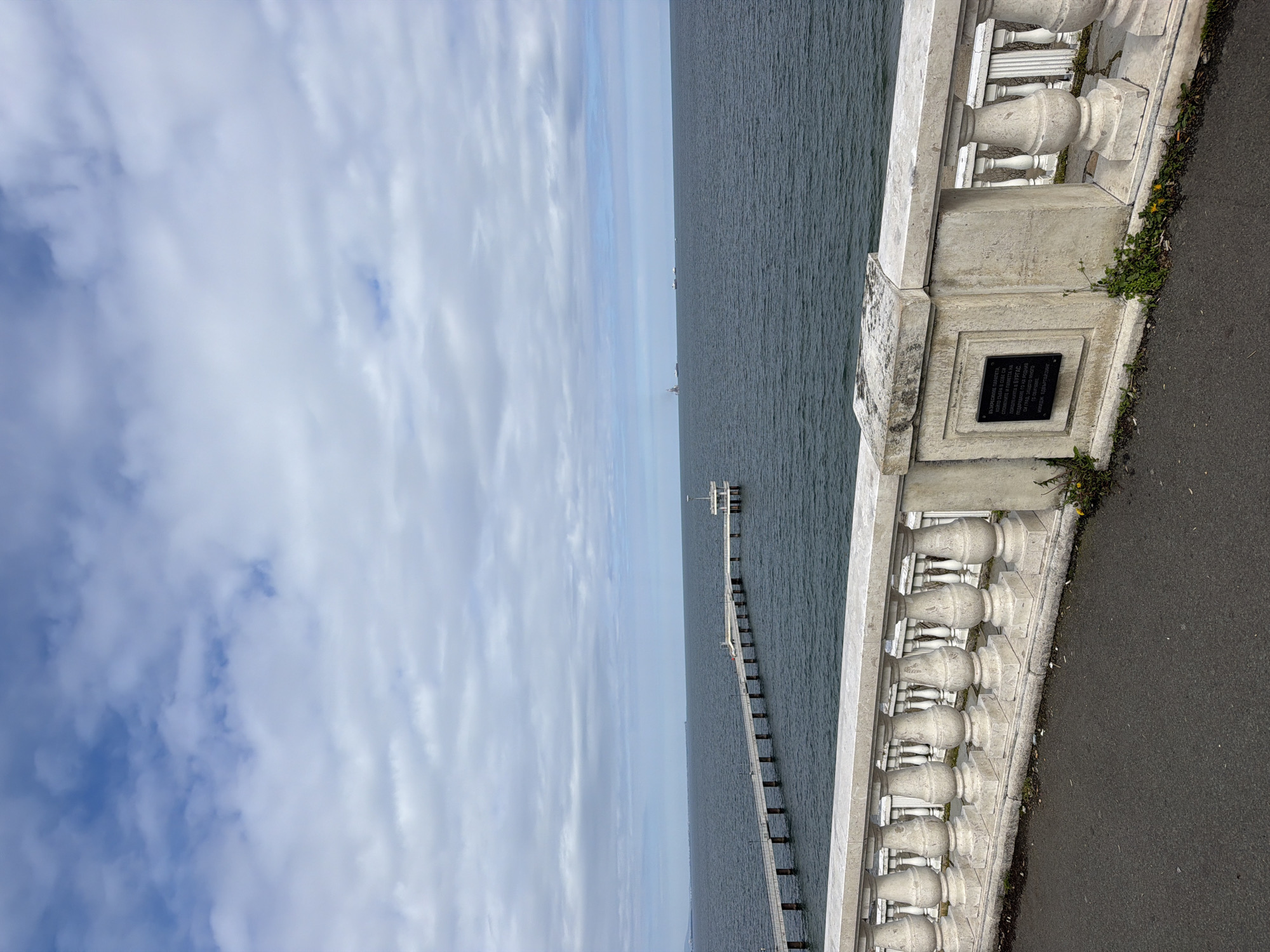 The image, taken in Burgas, Bulgaria, captures a serene coastal view under a partly cloudy sky. The perspective is from a paved promenade or pathway, looking out over the Black Sea.
In the right foreground, a white, ornate stone balustrade with intricately carved balusters runs horizontally. It separates the viewing area from the water. A dark rectangular plaque with white Cyrillic text is embedded into the balustrade's base. Below the balustrade, a thin strip of asphalt path is visible, with small weeds and yellow flowers growing along its edge.
Stretching from the mid-ground towards the distant horizon is a long, narrow pier, supported by numerous pilings. The pier extends far into the calm, dark blue-grey sea, with a structure or platform visible at its far end. Several faint, dark shapes, possibly ships or distant features, can be discerned on the horizon line.
The sky, occupying the left side of the frame, is a mix of light blue and scattered white, wispy clouds, suggesting an overcast or partly cloudy day. The overall scene conveys a peaceful, open coastal ambiance.
The text on the plaque, in Bulgarian Cyrillic, reads: "ВЪЗКРЕСЕНИ ПАРАПЕТА / КОЙТО СЪБРА В СЕБЕ СИ / СПОМЕНИТЕ И ПАМЕТТА НА / ПОКОЛЕНИЯТА В БУРГАС. / ПОДАРЯВАМЕ ГО НА РОДНИЯ / СИ ГРАД, ЗАЩОТО МНОГО / ГО ОБИЧАМЕ / ИМИДЖ ЕДВЪРТАЙЗИНГ". This translates approximately to: "Restored Balustrade, which gathered within itself the memories and remembrance of generations in Burgas. We gift it to our hometown because we love it very much. Image Advertising." This text highlights the local significance and sentimental value of the balustrade to the community of Burgas. No people or active events are visible, emphasizing the quiet beauty of the coastal landscape.