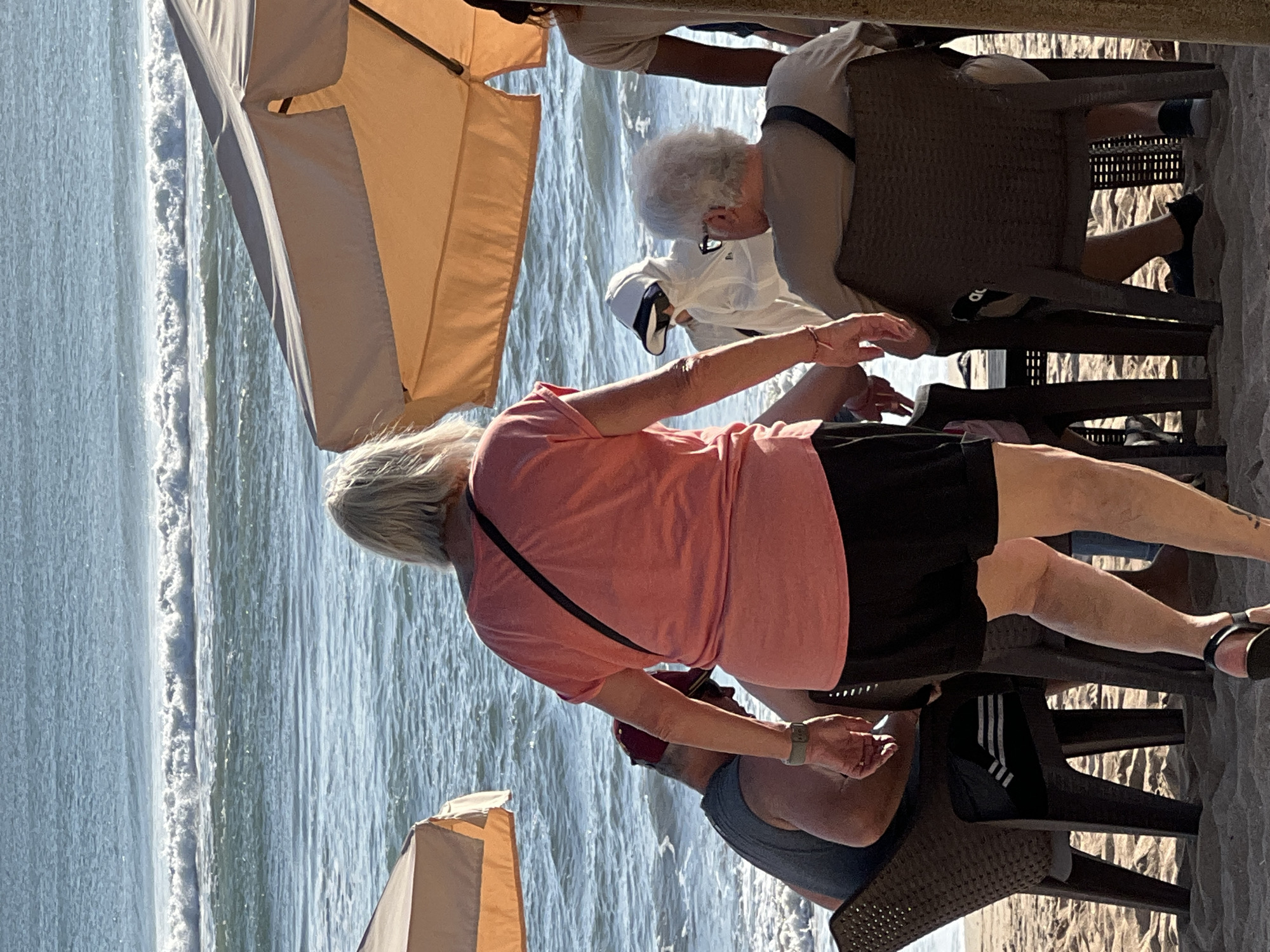 This image depicts a beach scene in Kuta, Indonesia. The primary focus is on a group of people seated at tables on the sand, with the ocean and waves visible in the background. A woman in a coral-colored t-shirt and black shorts is walking away from the camera, towards the water. To her left, a person wearing a grey tank top and a red baseball cap is seated. In front of them, under a beach umbrella, a person in a white hooded garment and sunglasses is visible. To the right of the woman in the coral shirt, another person with gray hair is seated, wearing a light-colored t-shirt. Further to the right, another person in a light-colored t-shirt is standing, partially obscured by an umbrella. The sand is light brown, and the water is a vibrant blue with white-capped waves. Beach umbrellas provide shade for the seated patrons.