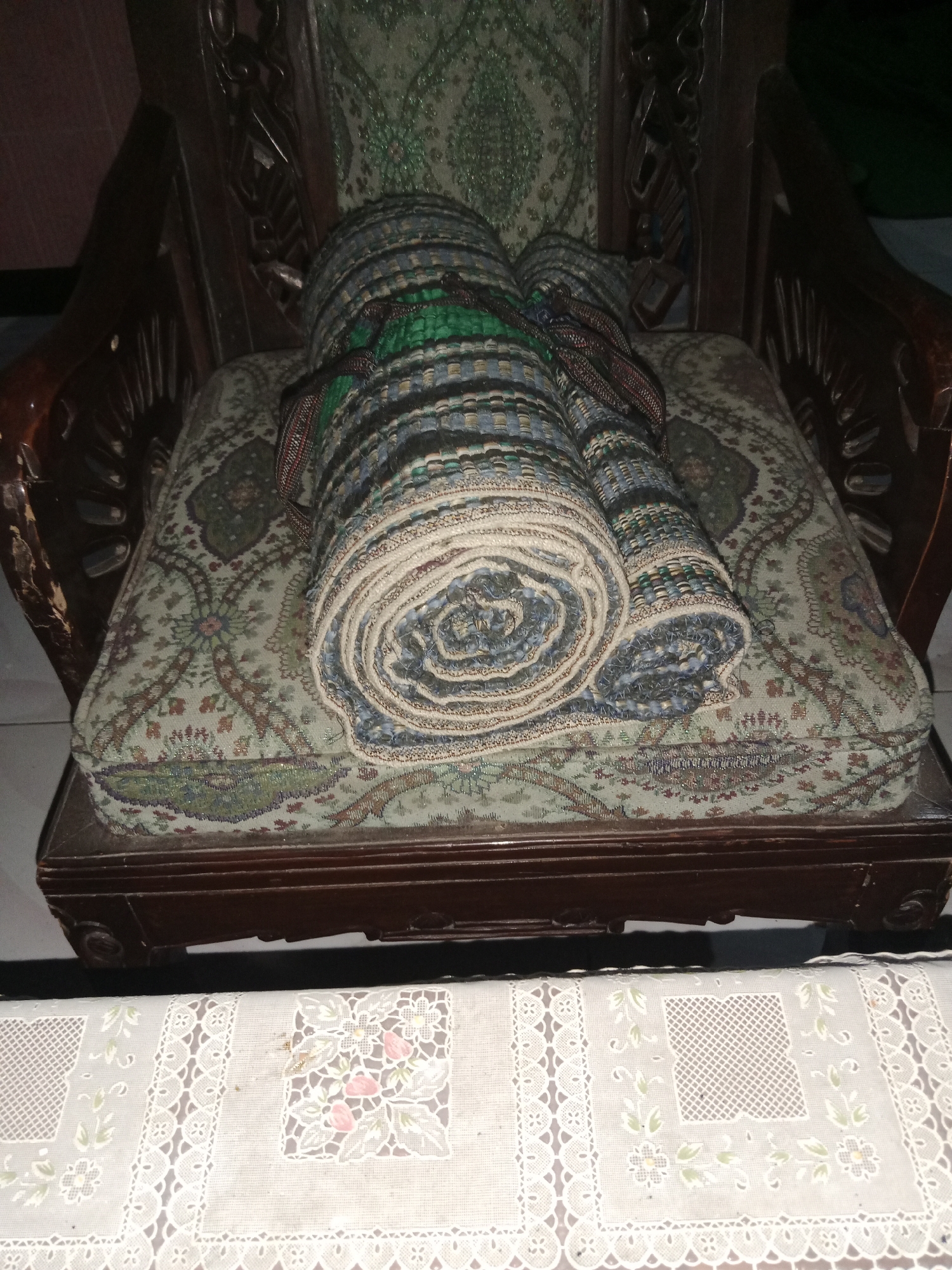 The image shows two rolled-up mats, possibly prayer mats or sleeping mats, resting on the patterned cushion of a dark wooden chair. The mats are tied together with a brown and red patterned strap. The chair itself has ornate carvings on its arms and back. In front of the chair, a white tablecloth with delicate lace trim and embroidered floral patterns is visible. The scene appears to be indoors, with a plain, dark background behind the chair, suggesting a residential setting. The location is identified as Ringinrejo, Indonesia.