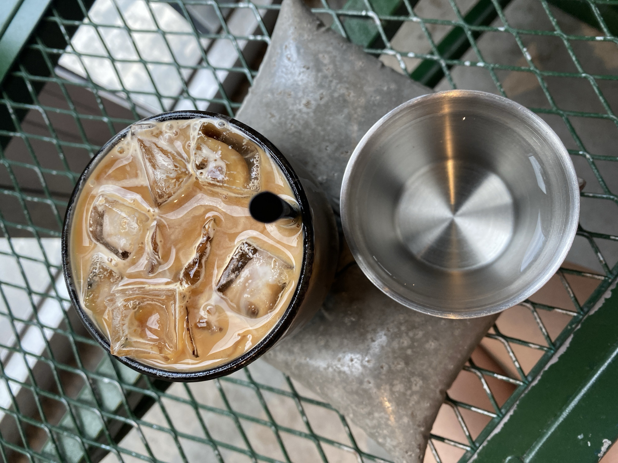 An overhead, high-angle shot shows an iced coffee drink and an empty metal cup on a green metal lattice table. The drink, in a dark rimmed glass, is filled with ice cubes and a creamy, light brown liquid. A black straw is visible in the drink. Next to the glass, an empty, shallow metal cup is placed with its concave side up. The table has a diamond pattern and appears to be outdoors, possibly at a cafe in Da Nang, Vietnam, given the style of outdoor furniture often found there. The background is blurred, showing the green lattice of the table and hints of the ground below.