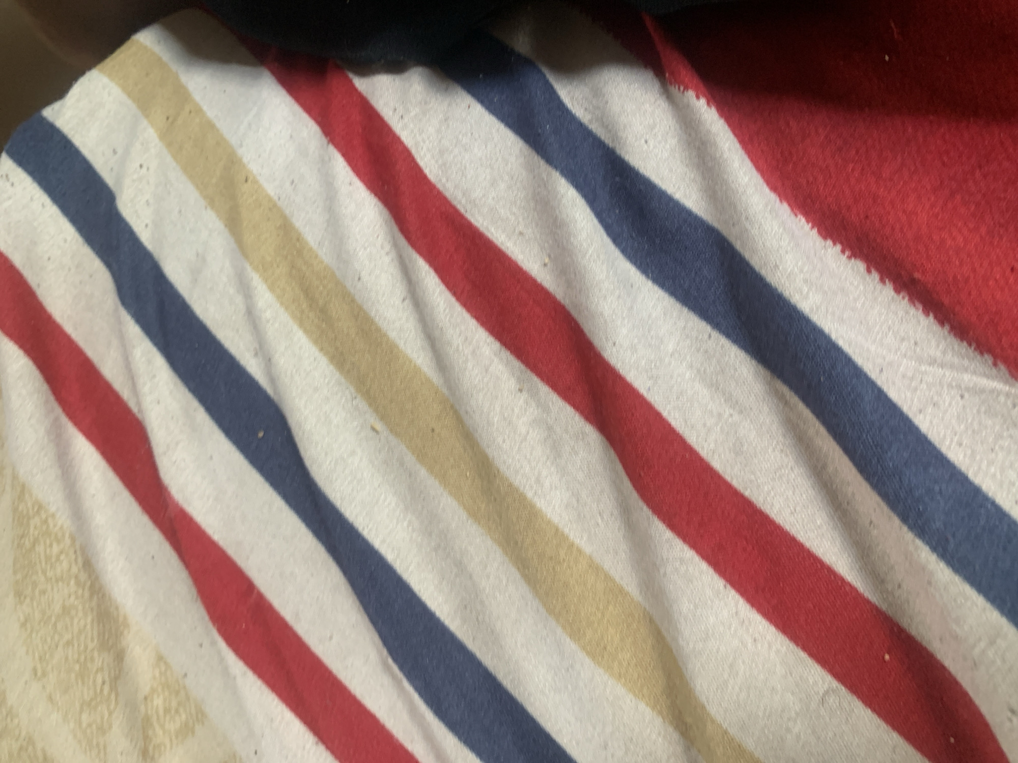 The image is a close-up, overhead shot of a striped fabric. The stripes run diagonally across the frame, alternating between red, white, blue, and a light yellow or beige. The fabric appears to be a blanket or throw, possibly cotton, with some visible texture and a few small specks of dirt or lint. In the upper left corner, there is a textured, light-colored surface, possibly a wall or another fabric. To the right, a dark, out-of-focus area suggests a shadow or another object. The scene is static and there are no people or discernible actions. The image does not provide any information that would allow for identification of its location in Jauro Yinu, Nigeria.