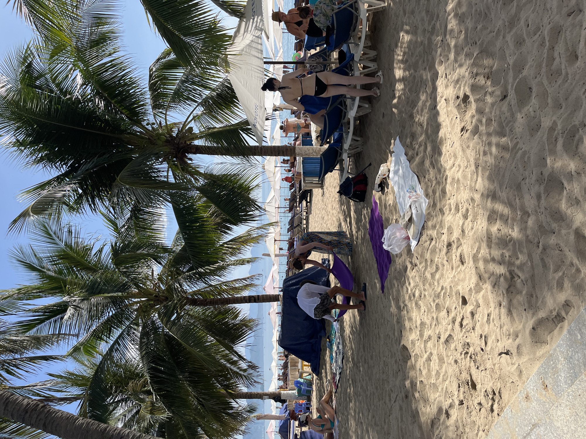This is a medium-shot, eye-level, outdoor photograph of a busy beach in Nha Trang, Vietnam. The beach is lined with palm trees, and in the background, there are many beach umbrellas and lounge chairs. Several people are visible on the sand, some relaxing, others interacting with each other.
In the foreground, two people are folding a purple cloth or towel. To their left, a woman in a blue bikini is seated on the sand. Further to the right, a woman in a black bikini is standing near lounge chairs, and another woman in a black top and white shorts is sitting in a chair. There are several beach towels and bags scattered on the sand. In the middle ground, a large blue tarp is set up, possibly as shade or for storage.
The sand is light brown and appears textured with footprints. The water is visible in the background, with distant hills or mountains beyond. The sky is clear and blue. The overall impression is of a sunny day at a popular tourist destination.