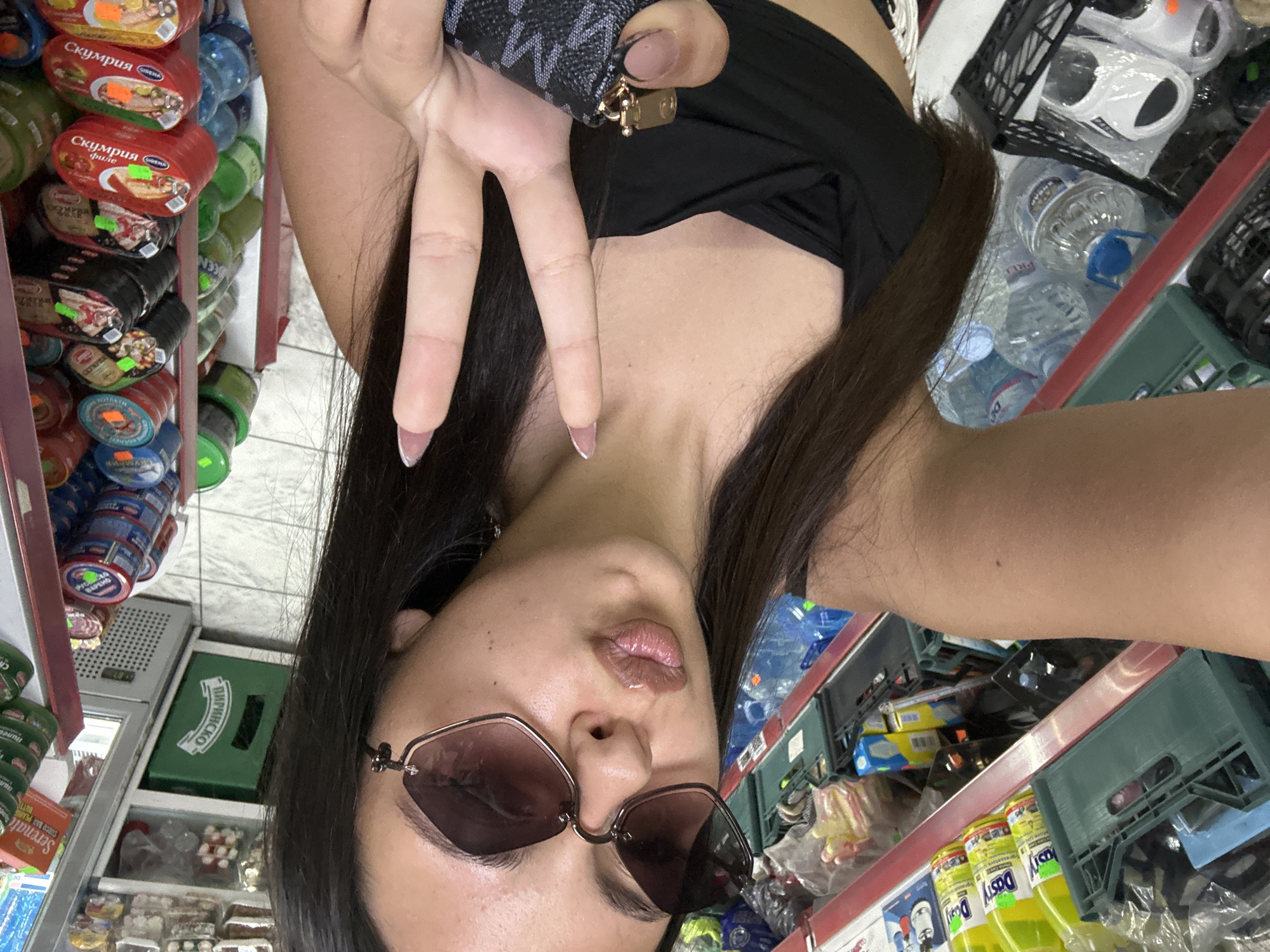 A person is seen taking a selfie in what appears to be a small grocery or convenience store. The image is rotated 90 degrees counter-clockwise. The subject is a female with long dark hair, wearing a black top and dark hexagonal sunglasses. She holds a small, dark patterned wallet or bag in her right hand while making a peace sign with her left hand, showcasing her manicured nails. Her lips are slightly puckered in a pout.
The background on the left side of the frame features shelves stocked with various canned goods. Several red cans are visible, with Cyrillic text, including "Скумрия филе" (Mackerel fillet) and "Скумрия" (Mackerel). Price tags are attached to some of the cans. Further down, blue and green cans are also present.
On the right side of the frame, there are shelves or crates holding large bottles of water, and other packaged items. A prominent green crate with "ПИРИНСКО" (likely "Pirinsko," a Bulgarian beer brand) is visible. Below, yellow bottles labeled "Dasty" are seen, alongside a package that reads "Serenat CHOCO BAR PEANUT BUTTER." The overall setting suggests a retail environment with various food and beverage products. The lighting indicates an indoor setting.