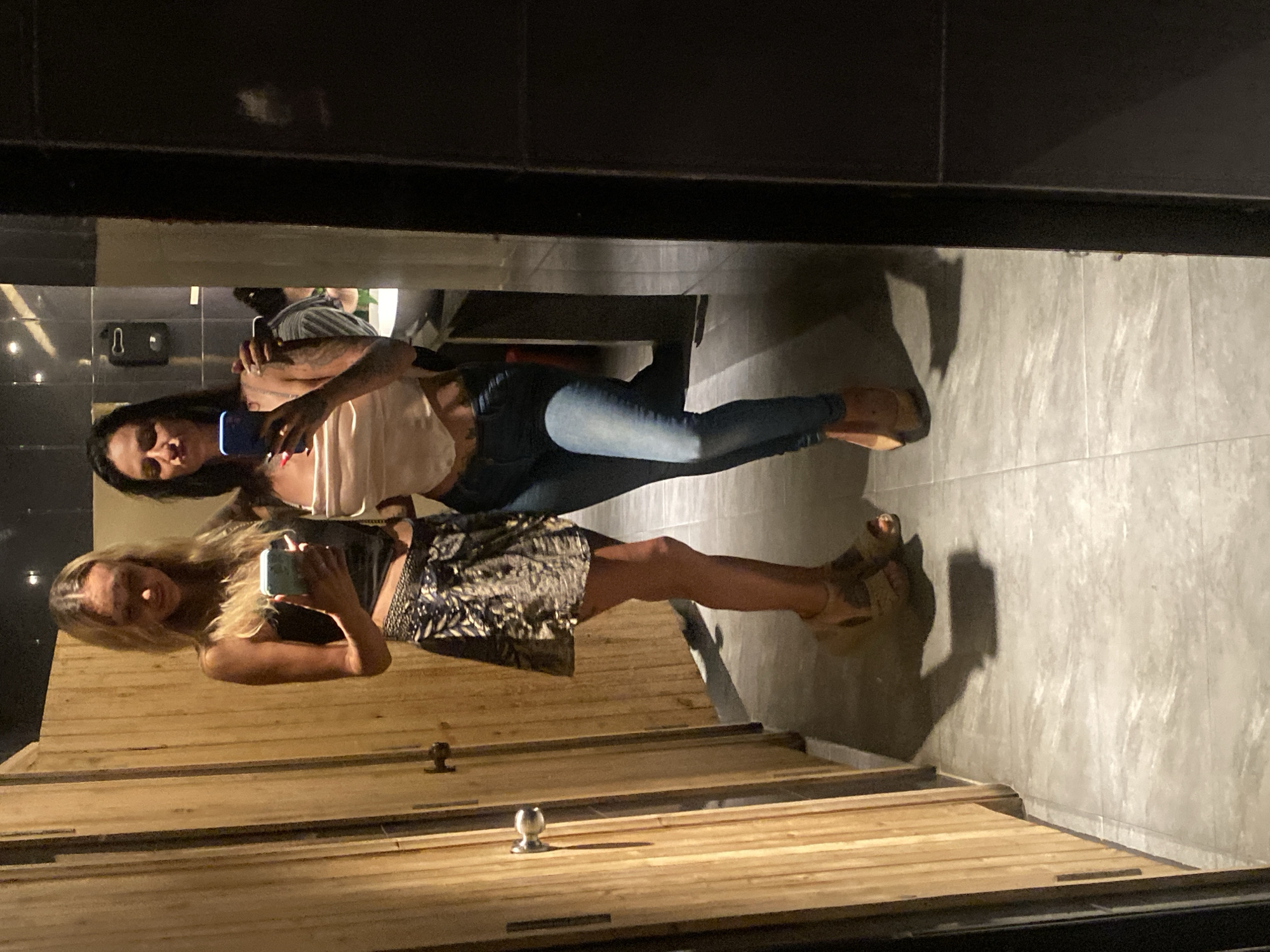 Two women are reflected in a mirror. The woman on the left has long blonde hair, is wearing a black top, and a patterned skirt. She is holding a blue phone. The woman on the right has dark hair, is wearing a light-colored top, and blue jeans. She is holding a purple phone. The reflection shows a bathroom or restroom with wooden stall doors and grey tile walls. The location is Phu Thuy Ward, Vietnam.