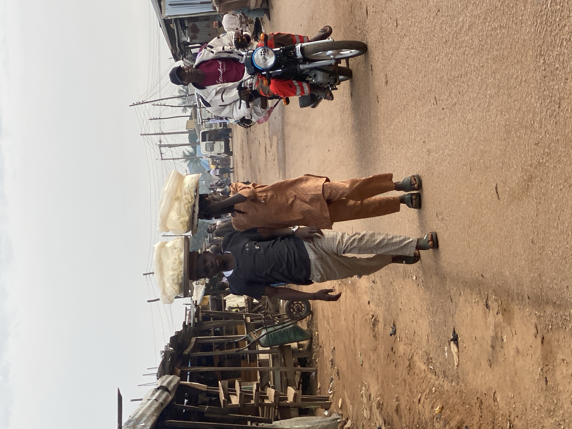 In Sabon Tasha, Nigeria, two young boys are walking side-by-side down a dirt road. The boy on the left wears a black t-shirt and plaid pants, while the boy on the right is dressed in a brown two-piece outfit. Both boys have large bundles of white material balanced on their heads. Behind them, a man on a motorcycle wearing a white jacket over a maroon shirt is riding towards the viewer. The road is lined with wooden stalls and some parked vehicles. The sky is overcast.