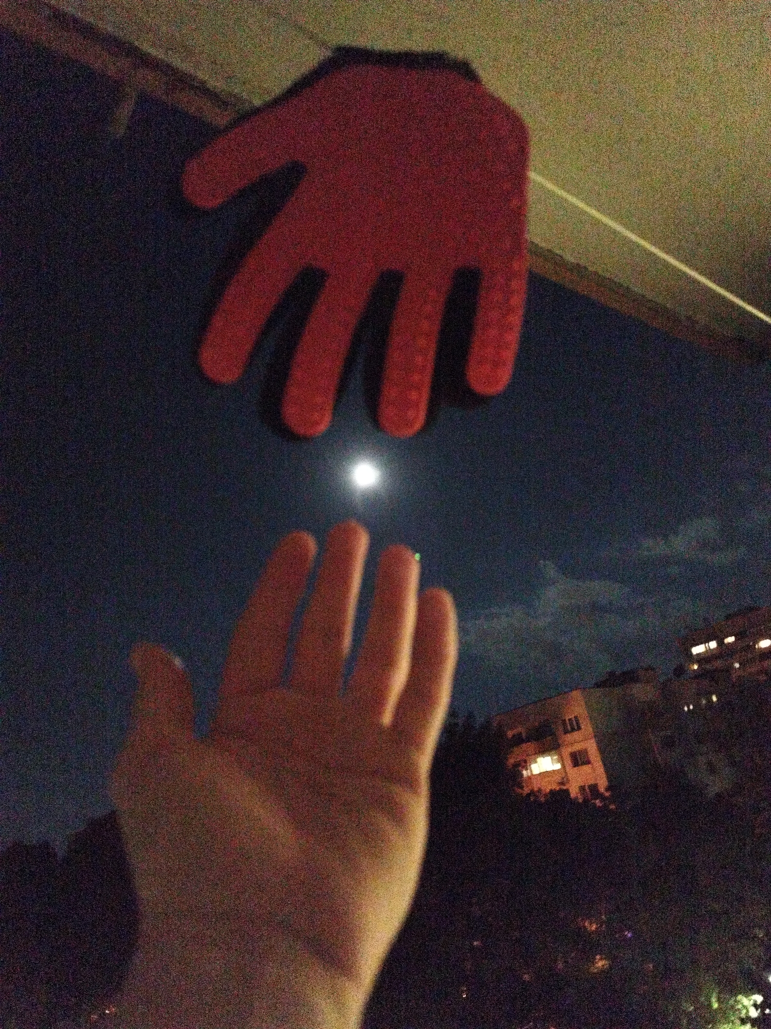 The image depicts a hand reaching towards the night sky, with a red hand-shaped object suspended above it. The background reveals a night scene with a visible moon and building silhouettes suggesting an urban environment. Buildings appear to be in the distance, potentially part of a residential area. There are visible clouds in the night sky. Based on the architectural style of the building in the background and the general topography, this could possibly be a neighborhood in Sofia, Bulgaria, but without more specific landmarks, this identification cannot be confirmed.