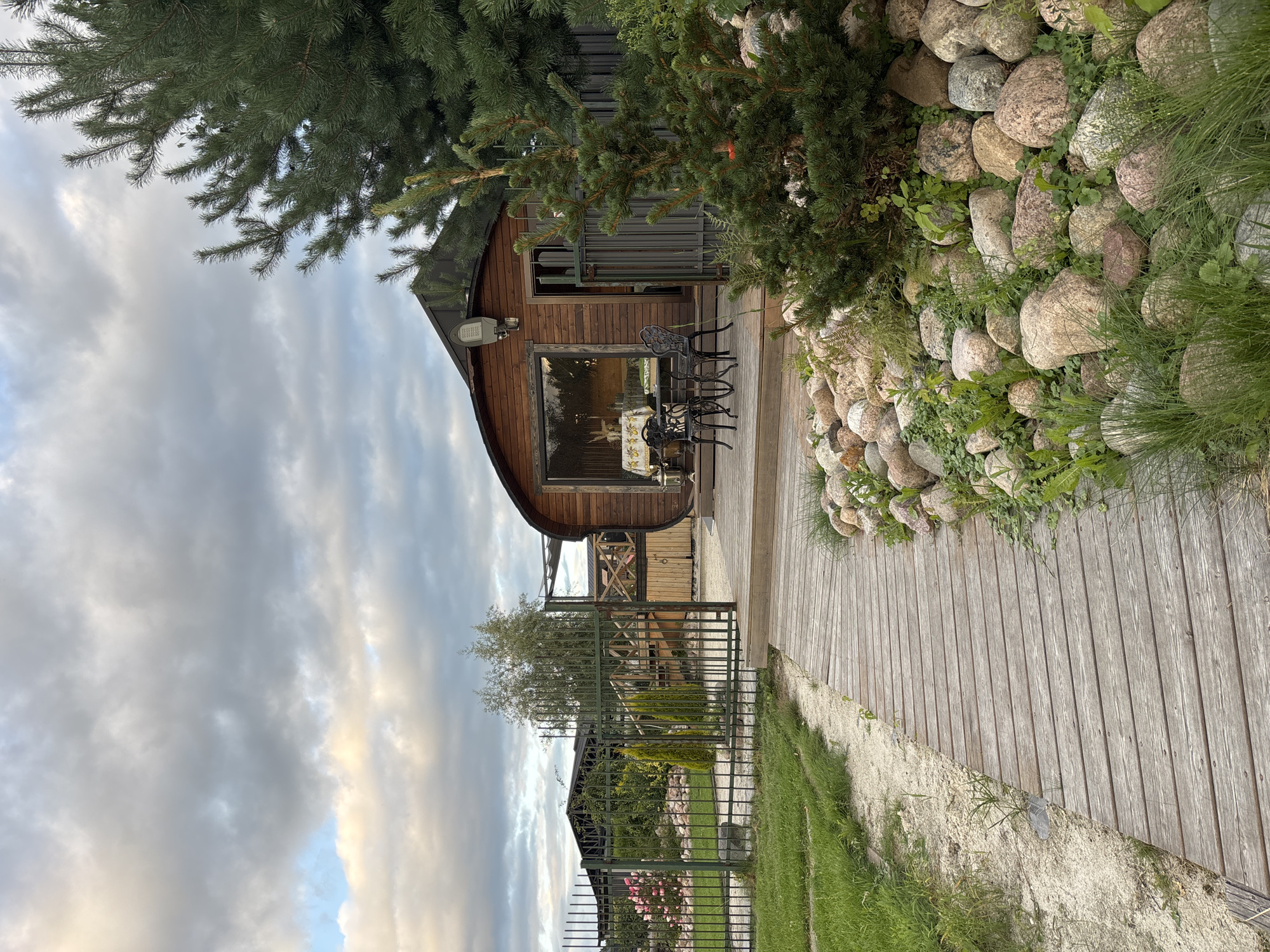 The image depicts a wooden boardwalk leading towards a barrel-shaped wooden building with a large window. Outside the building, on a wooden deck, are a table and chairs. The area surrounding the boardwalk and building is landscaped with rocks, grass, and a young pine tree in the foreground. A metal fence is visible, separating the property from a garden with rose bushes and other greenery. In the background, there are more structures and trees under a cloudy sky. The scene appears to be a resort or a private property, potentially located in Zvenigorod, Russia, given the context. There are no people or animals visible in the image.